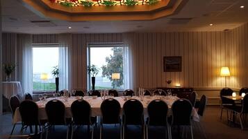 Un salón de conferencias con una mesa grande dispuesta para una reunión, dos ventanas con cortinas, una lámpara decorativa en el techo y una lámpara en la pared.
