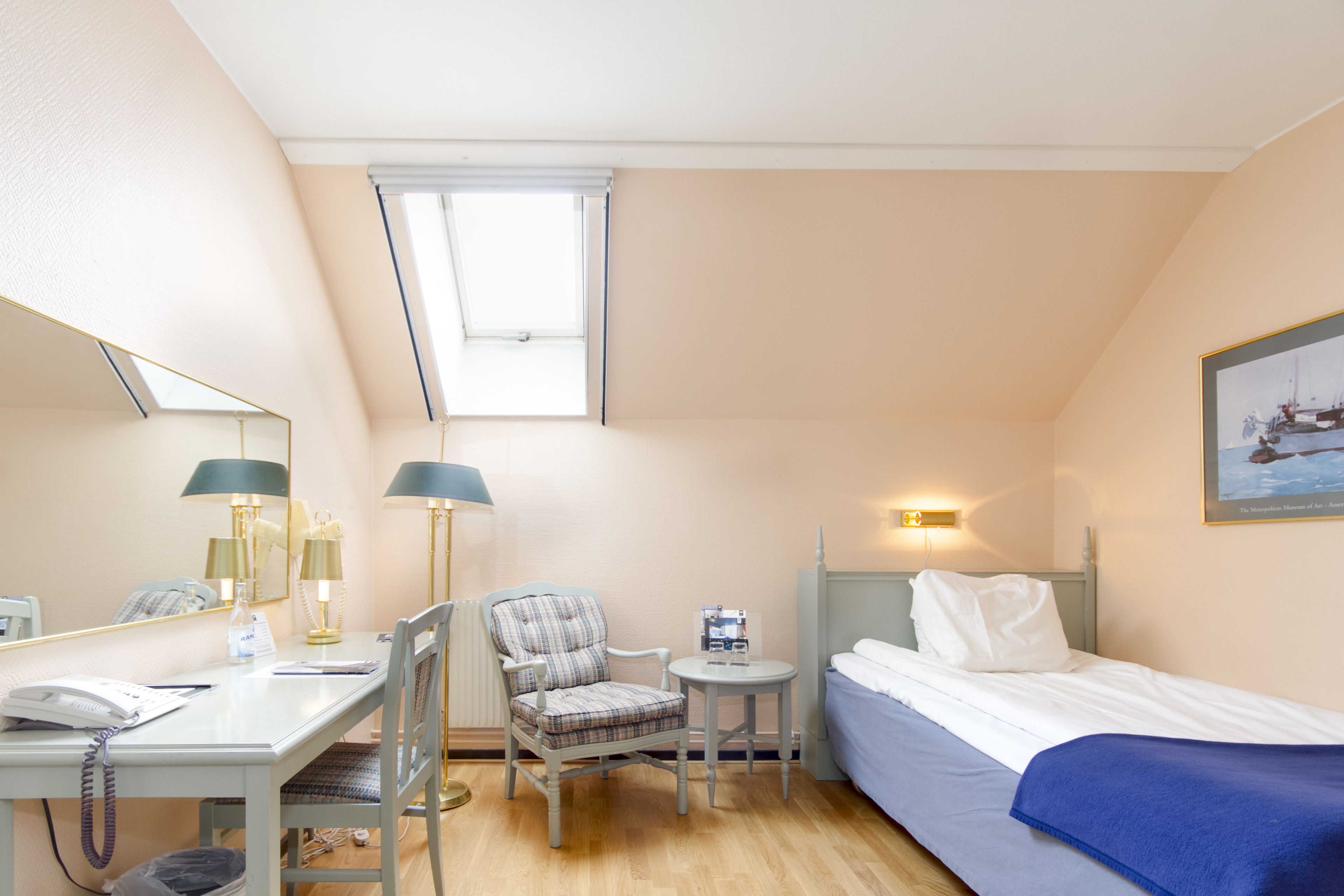 A hotel room with a bed, a desk with a telephone, a chair, a small table, and a lamp.