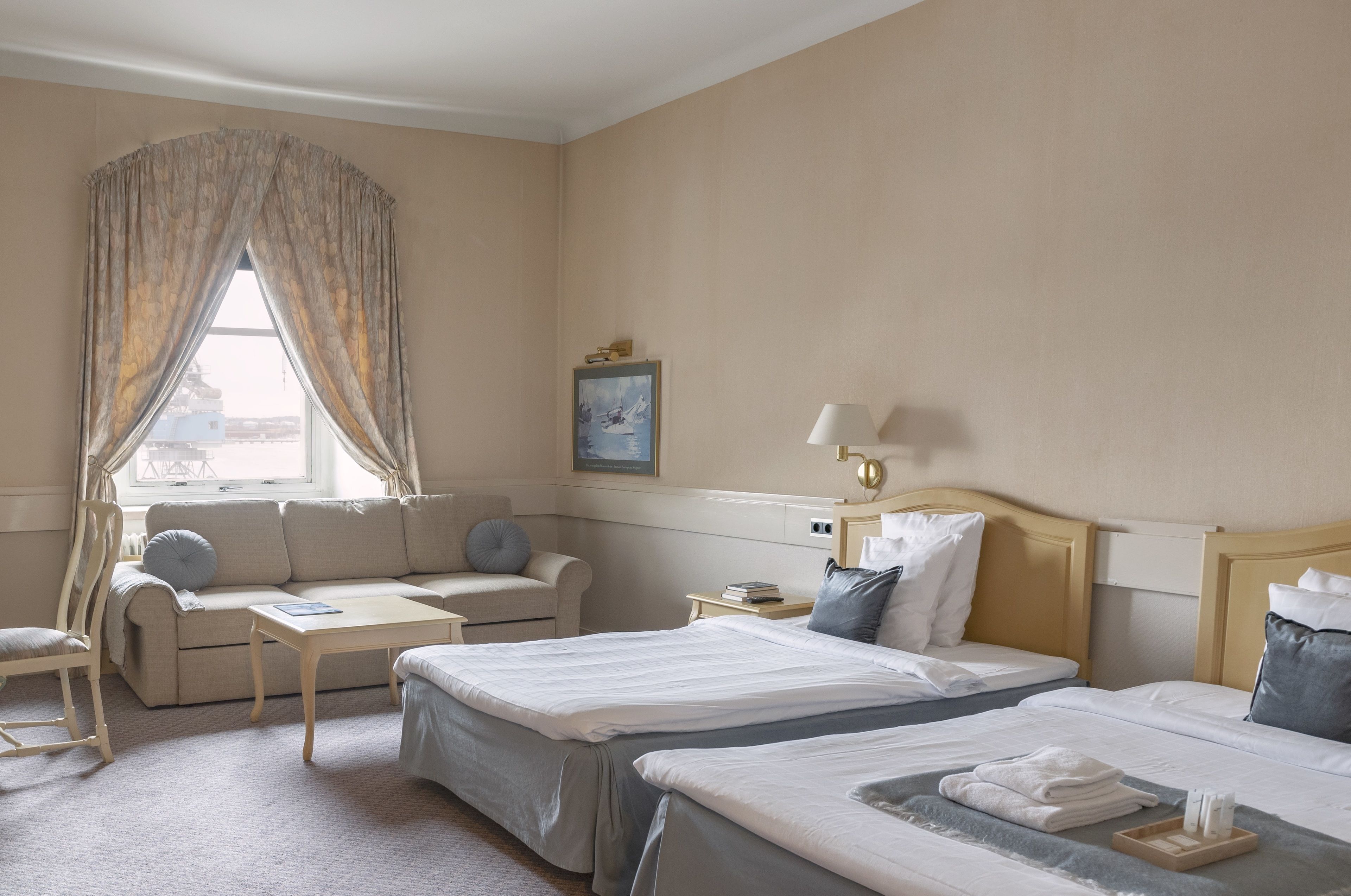 Habitación de hotel con dos camas, un sofá, una mesita y una ventana con cortinas.