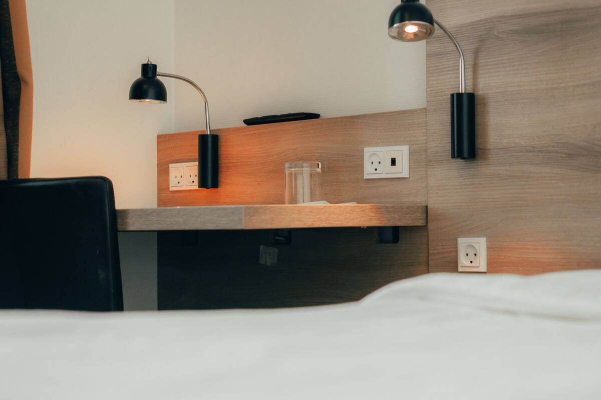 Habitación de hotel con cabecera de madera, un escritorio con silla, un vaso de agua y dos lámparas de pared.