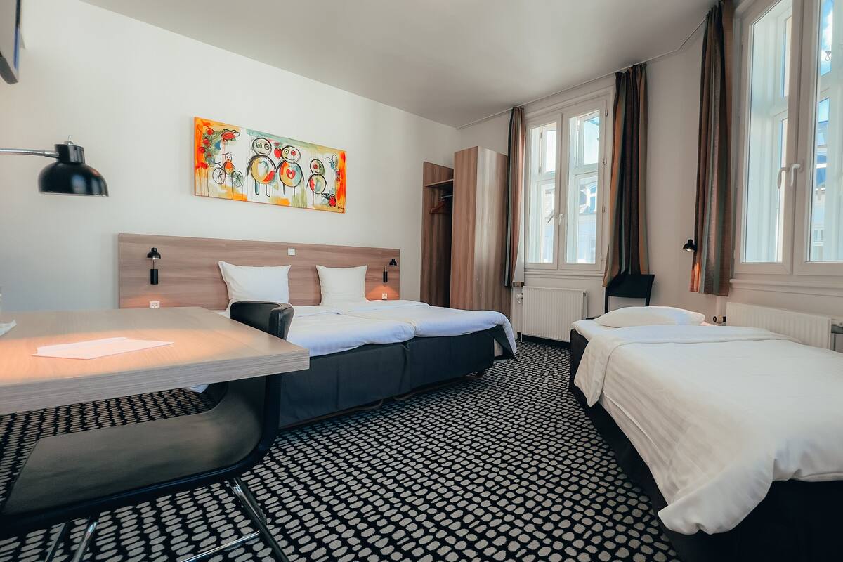 Habitación de hotel con dos camas, un escritorio, una silla, un cuadro en la pared y ventanas amplias con cortinas.