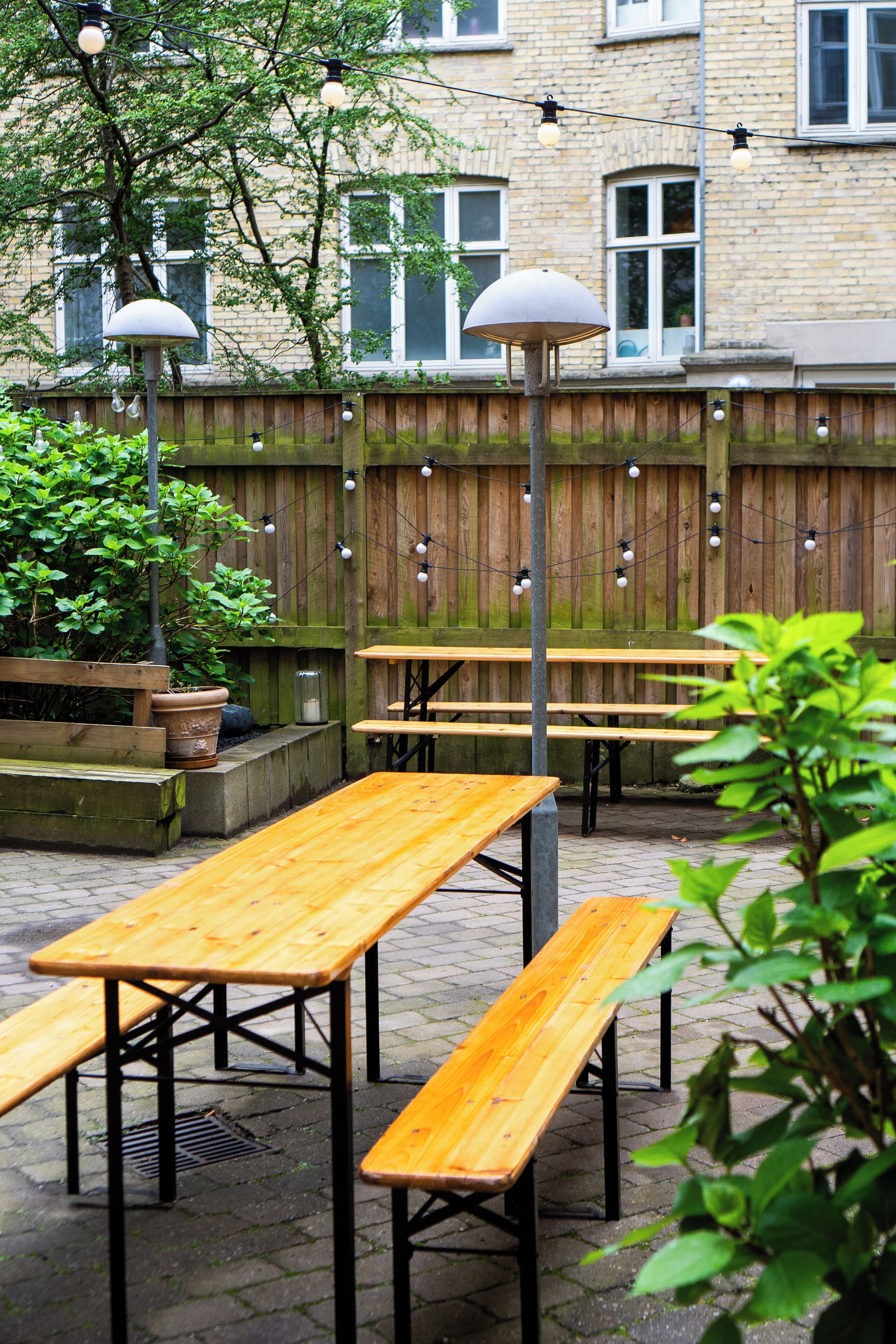 An outdoor patio area with wooden benches and tables, string lights, and a wooden fence.