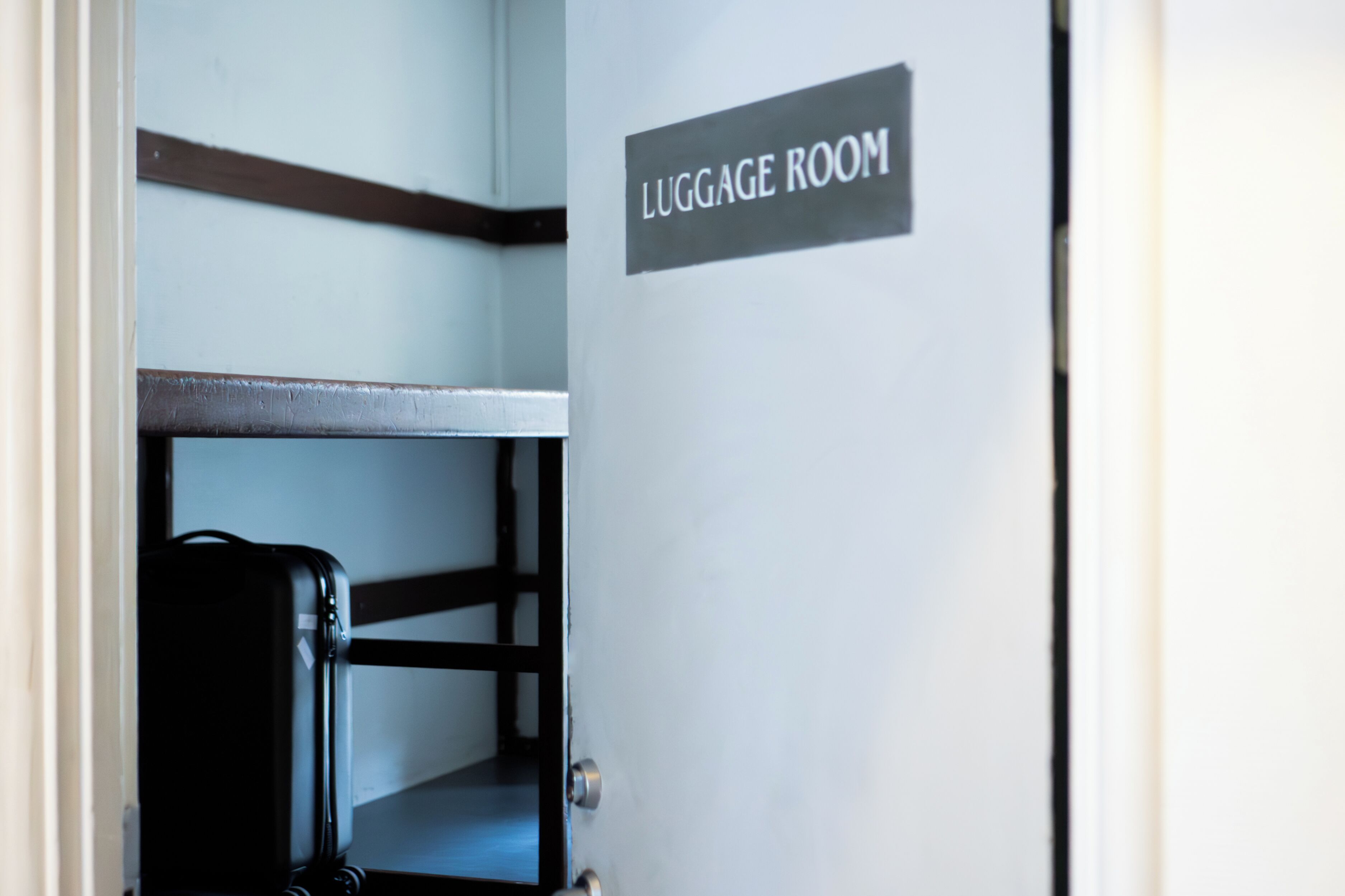 Een gesloten deur met een bord waarop 'LUGGAGE ROOM' staat, en een zwarte koffer op een metalen bank.