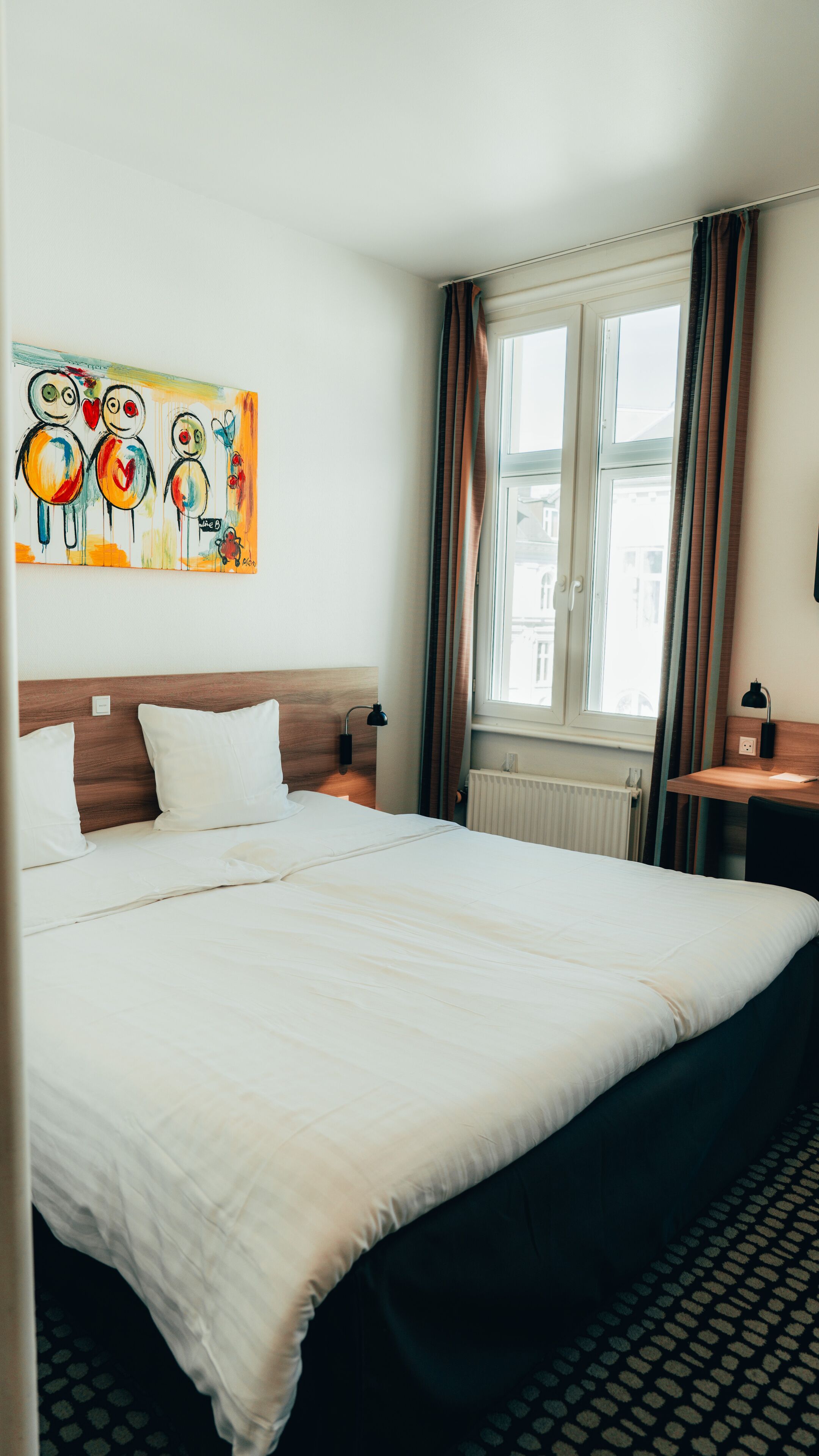 Een hotelkamer met een groot bed, een hoofdeinde, een nachtkastje met een lamp, een raam met gordijnen, en een kleurrijk schilderij aan de muur.
