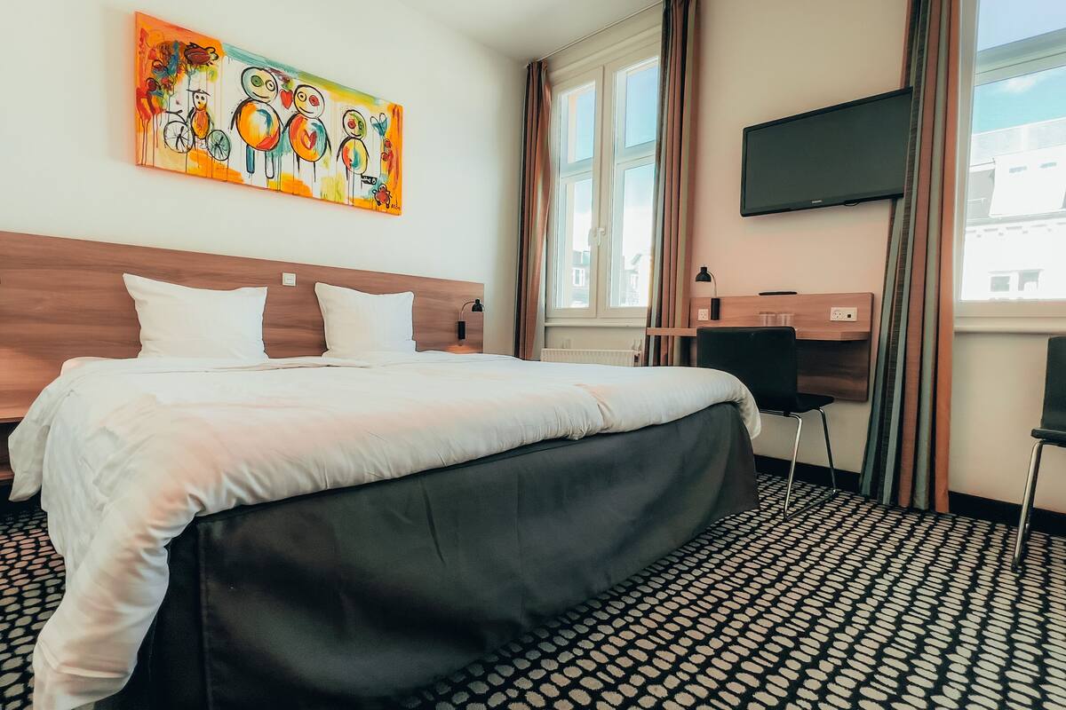 Habitación de hotel con una cama grande, un escritorio con silla, un televisor en la pared y una ventana con cortinas.