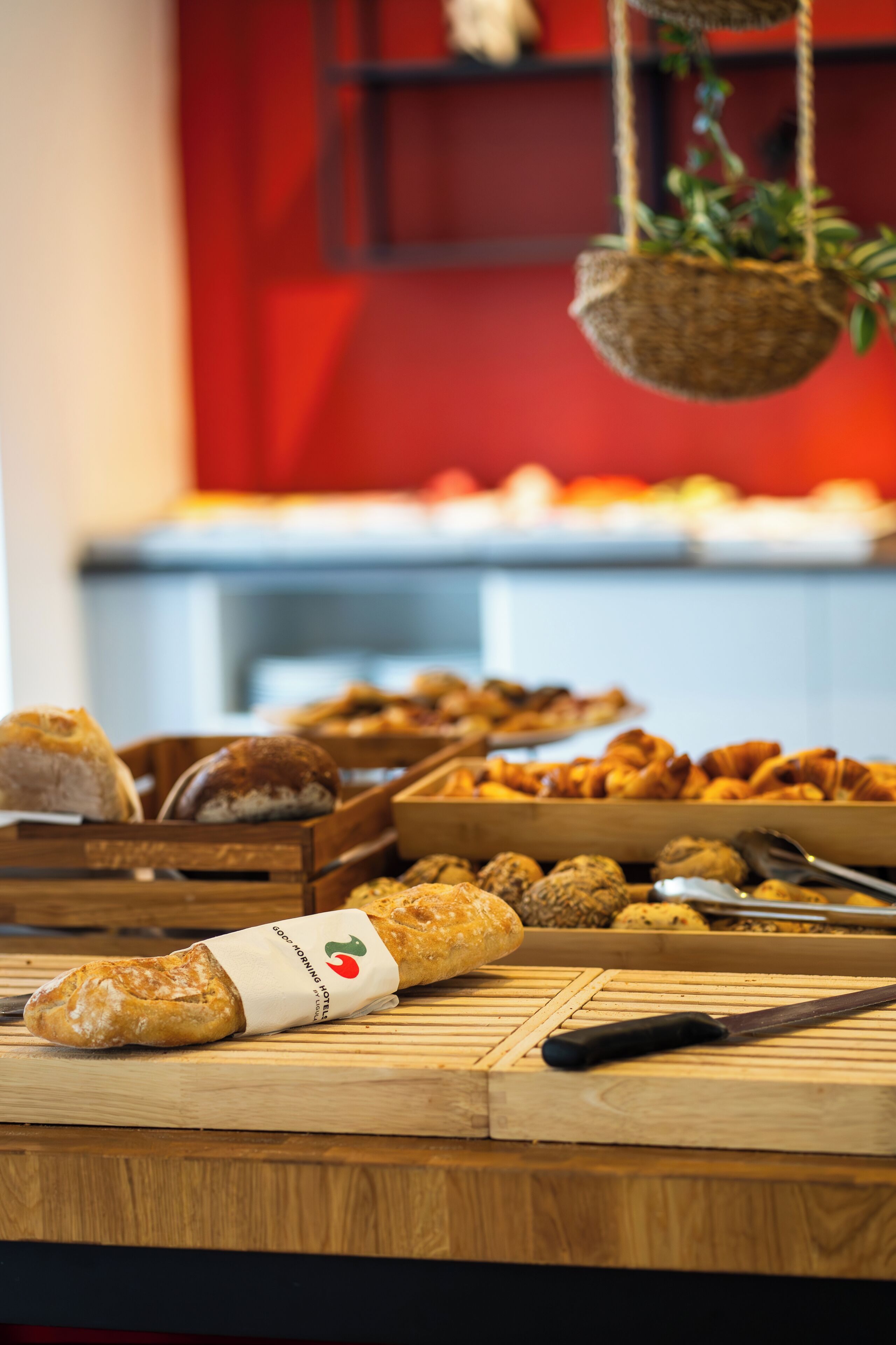 A wooden table with various baked goods, including bread and pastries, in a kitchen setting with a red wall and hanging plant.