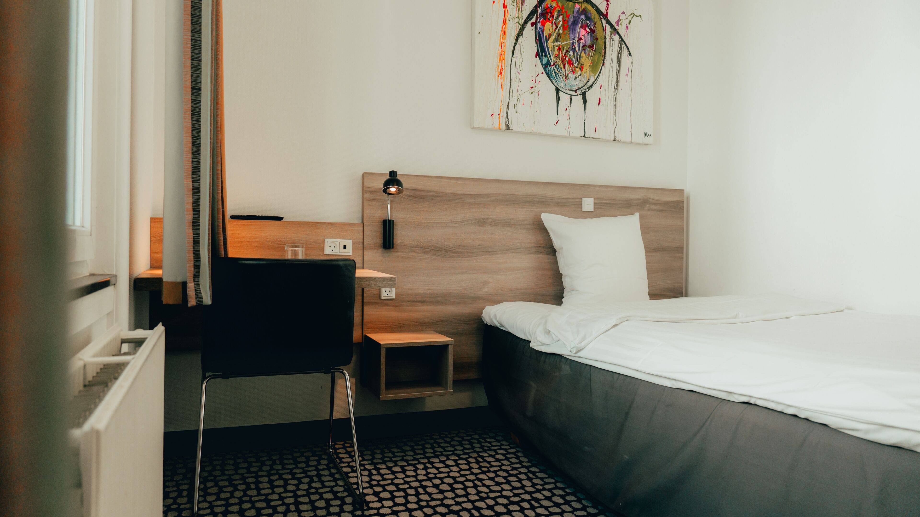 Una habitación de hotel con una cama, un escritorio, una silla, una mesita, una lámpara de pared y un cuadro en la pared.