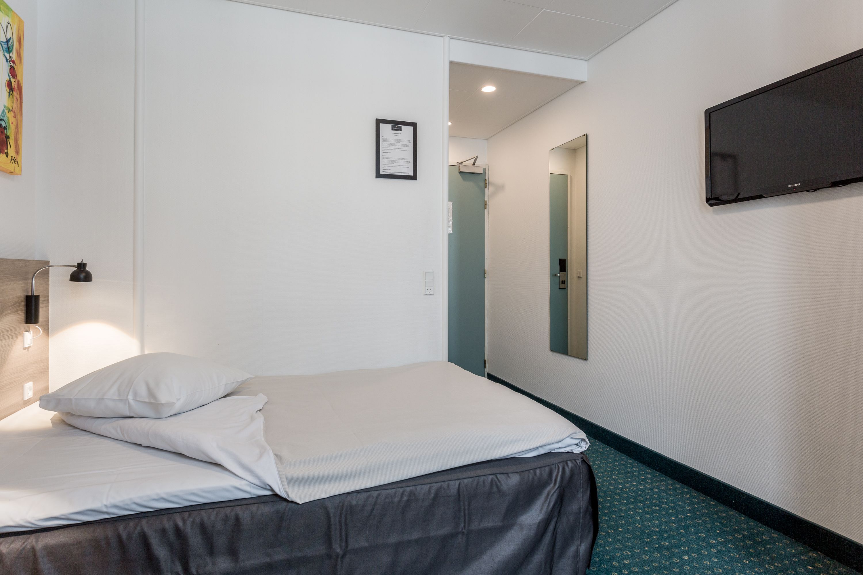 Habitación de hotel con cama, televisor de pantalla plana montado en la pared, un certificado enmarcado y una puerta que conduce a un baño.