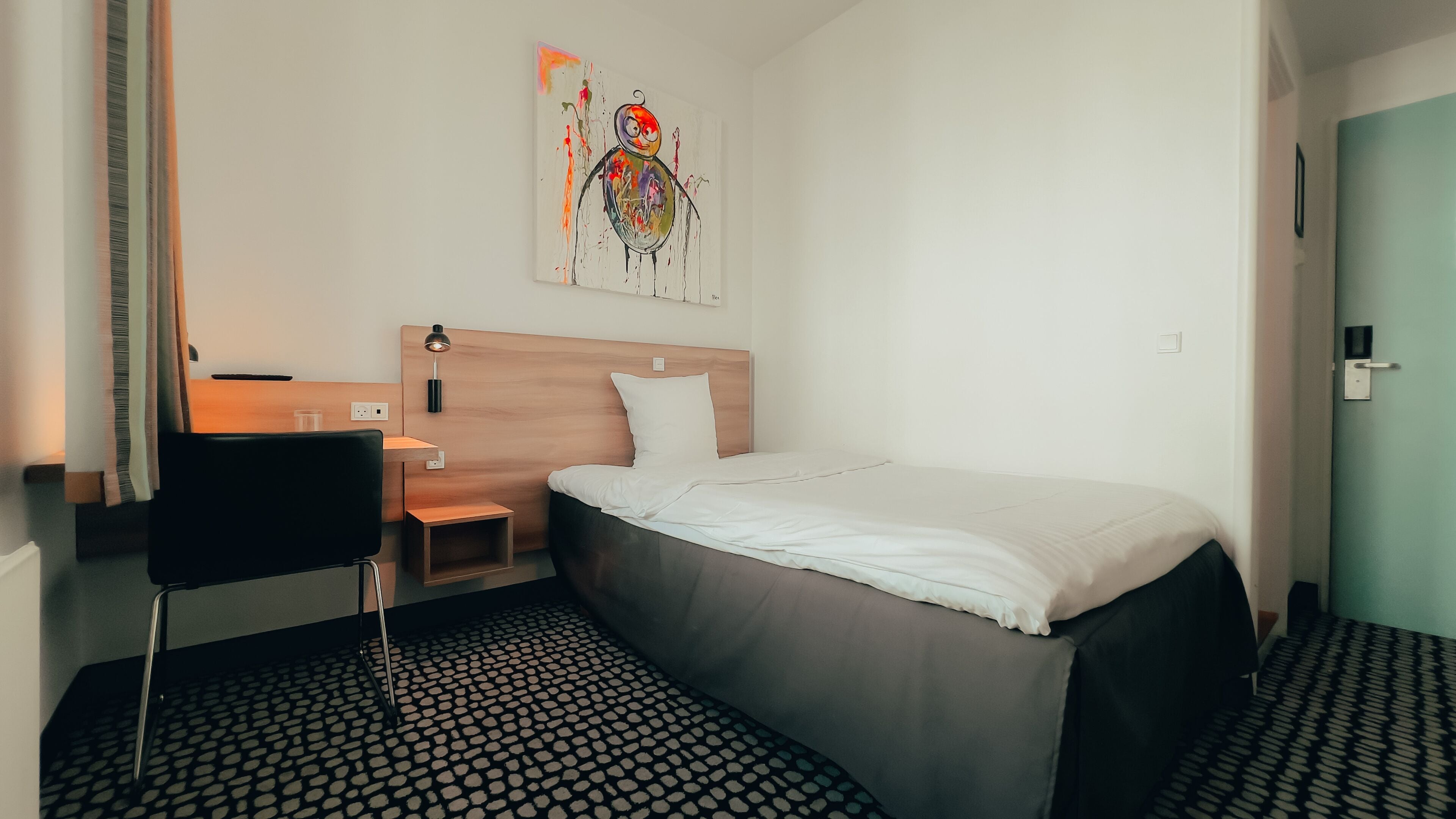 A hotel room with a bed, a desk with a chair, a small table, a wall-mounted lamp, and a colorful abstract painting.