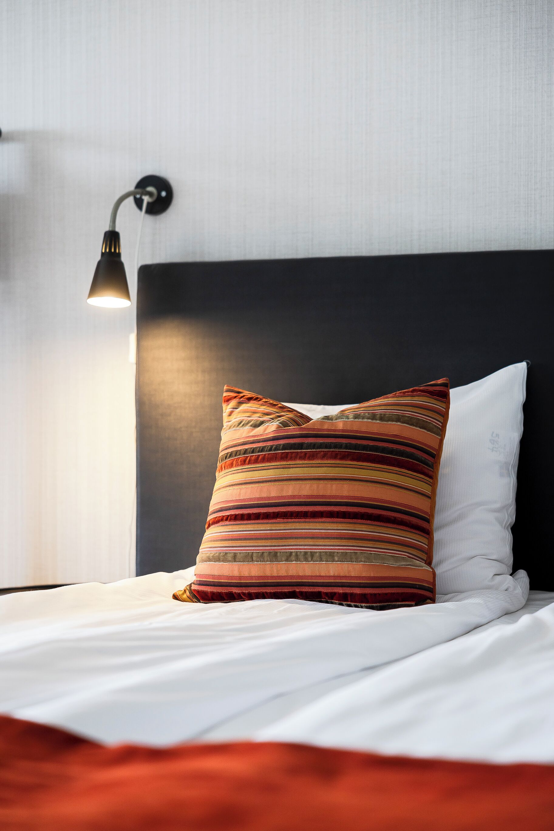 Una cama bien hecha con almohadas a rayas y lisas, un cabecero negro y una lámpara fijada a la pared.