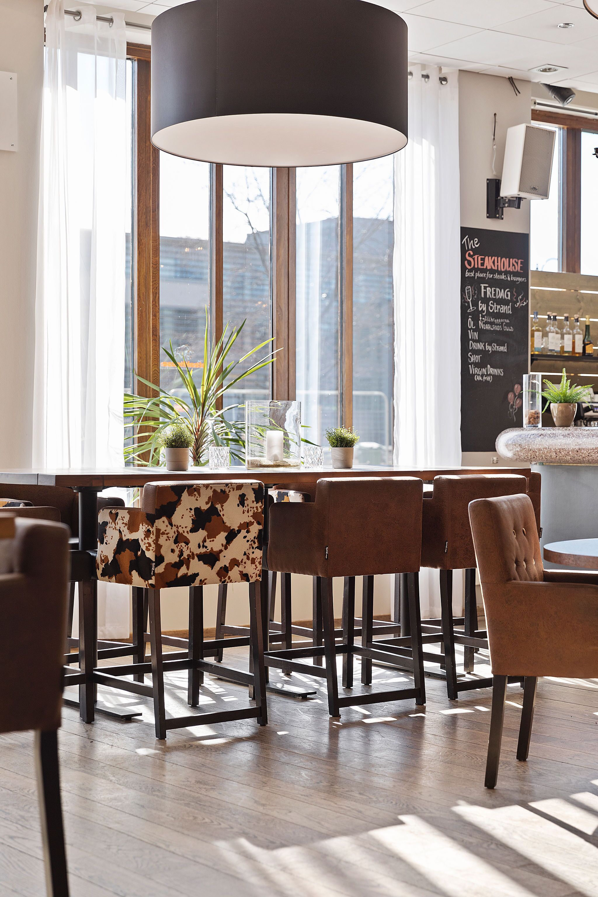Interior de un restaurante con una lámpara colgante negra grande, mesas de madera y sillas con estampado de cuero de vaca.