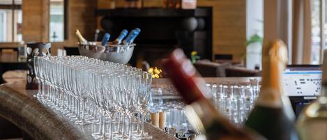 A bar area with multiple glasses lined up on a bar counter, a bottle of champagne, and a bowl of utensils in the background.