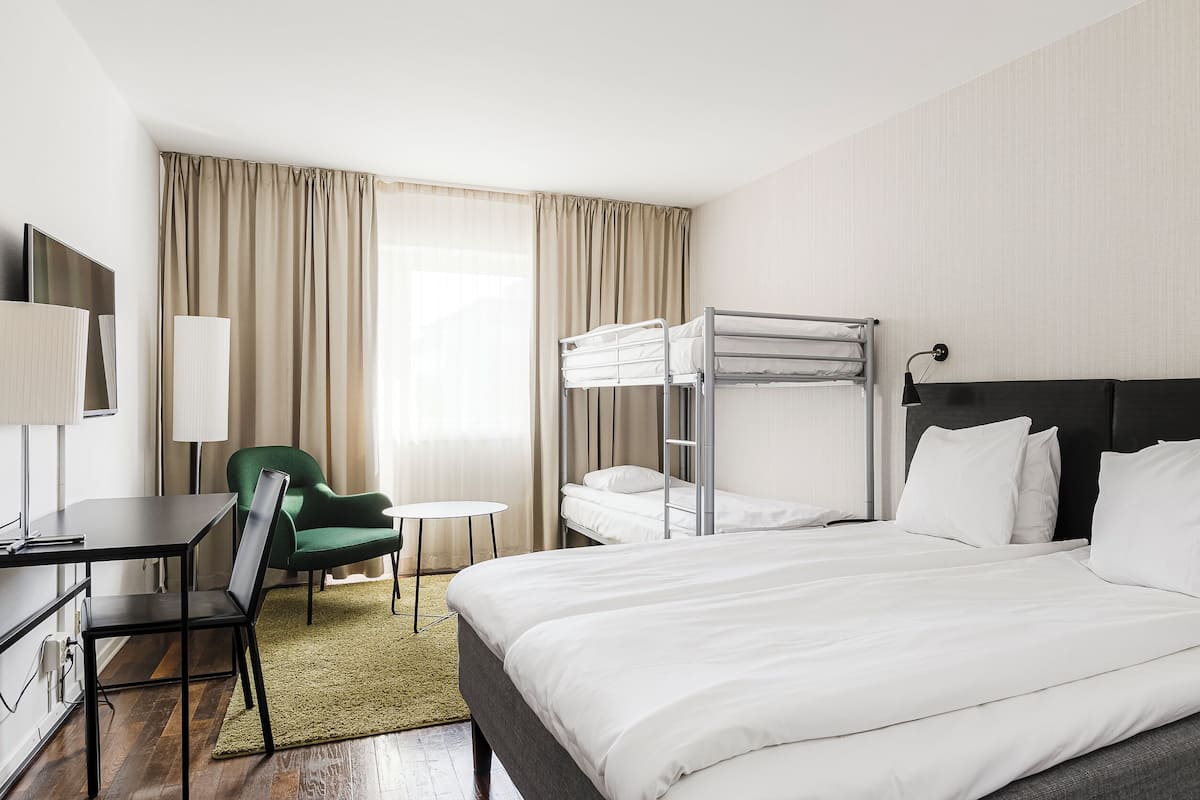 Une chambre d’hôtel avec un lit superposé, un bureau avec une chaise, une petite table ronde et un fauteuil vert.