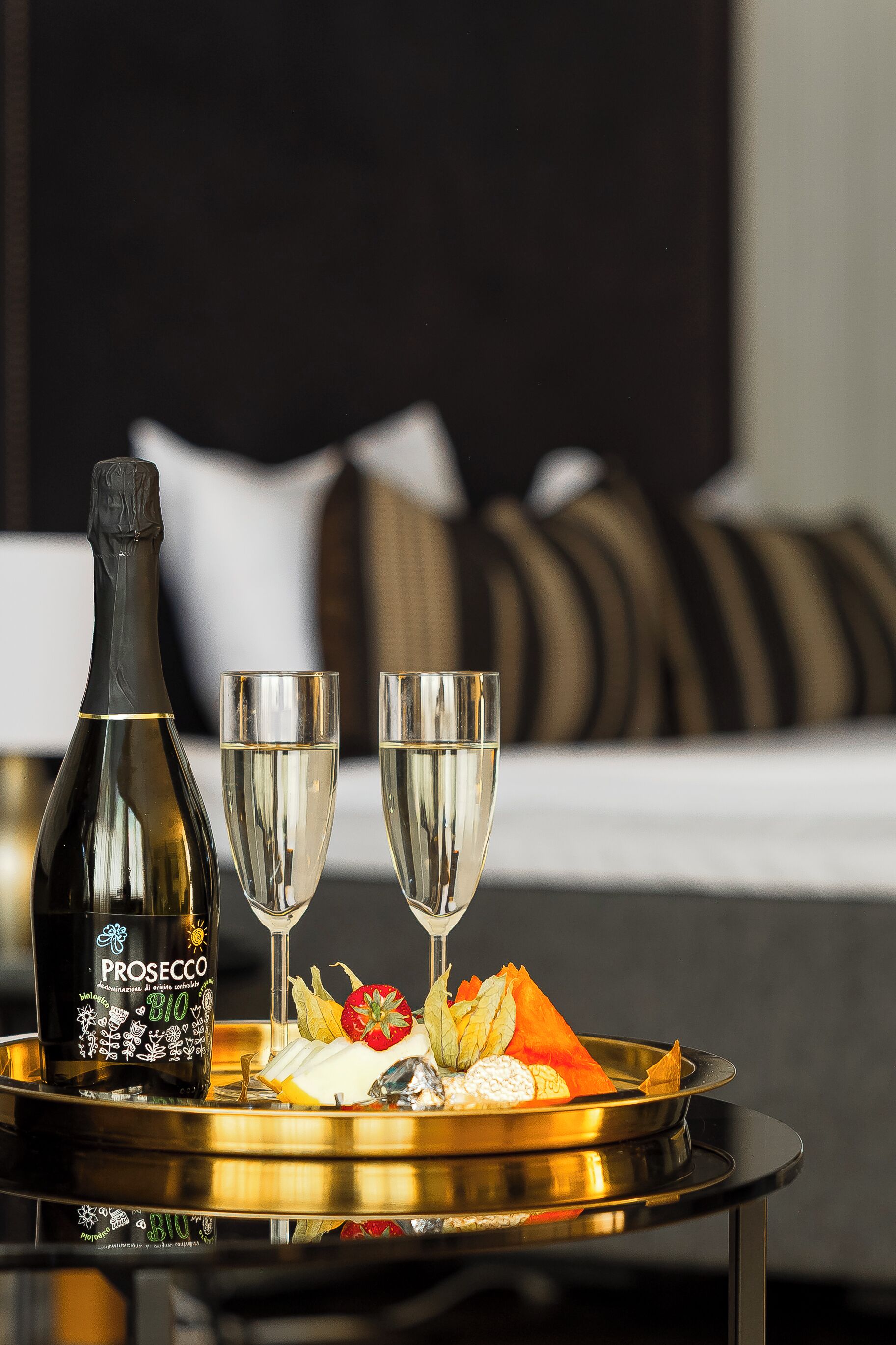 Uma garrafa de Prosecco, dois copos e um tabuleiro com comida e flores sobre uma mesa num quarto de hotel.