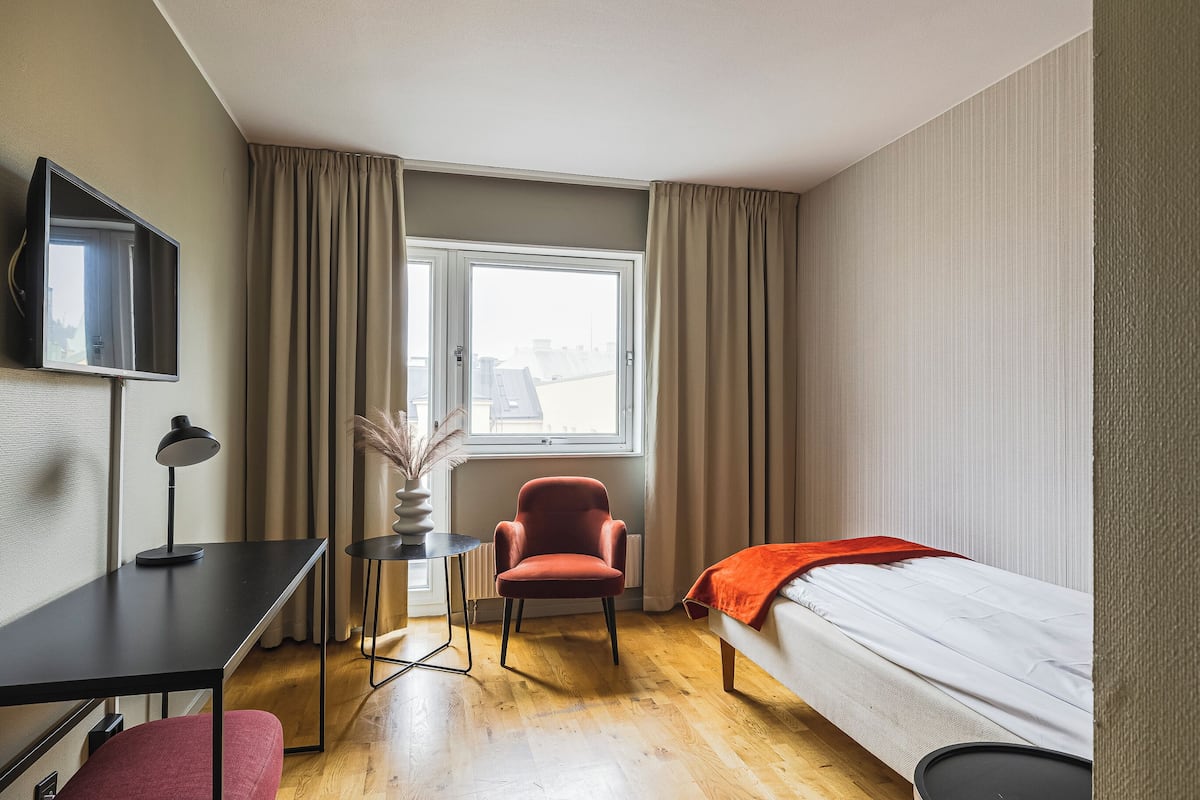 Une chambre d’hôtel avec un lit, un bureau avec une lampe, une chaise, un téléviseur fixé au mur et une fenêtre avec des rideaux.