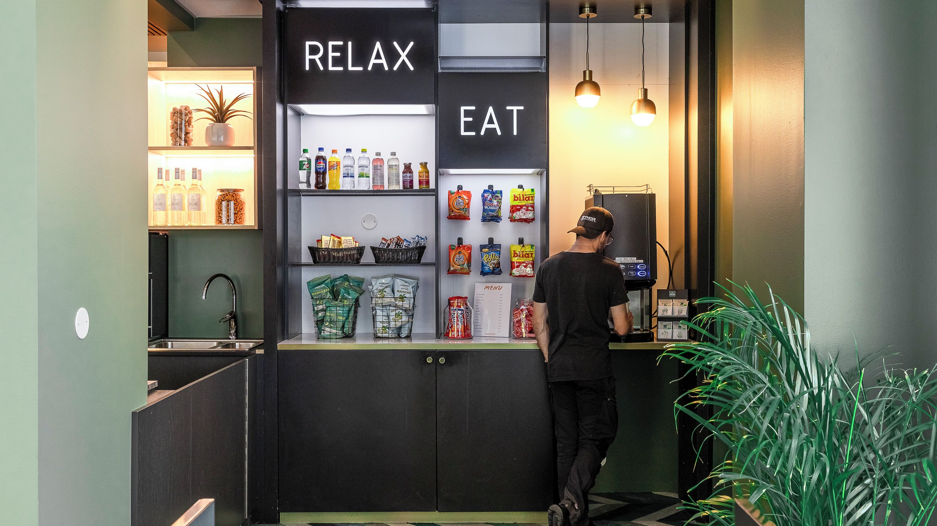 A self-service beverage station with a menu and snacks, a sink, and a potted plant.
