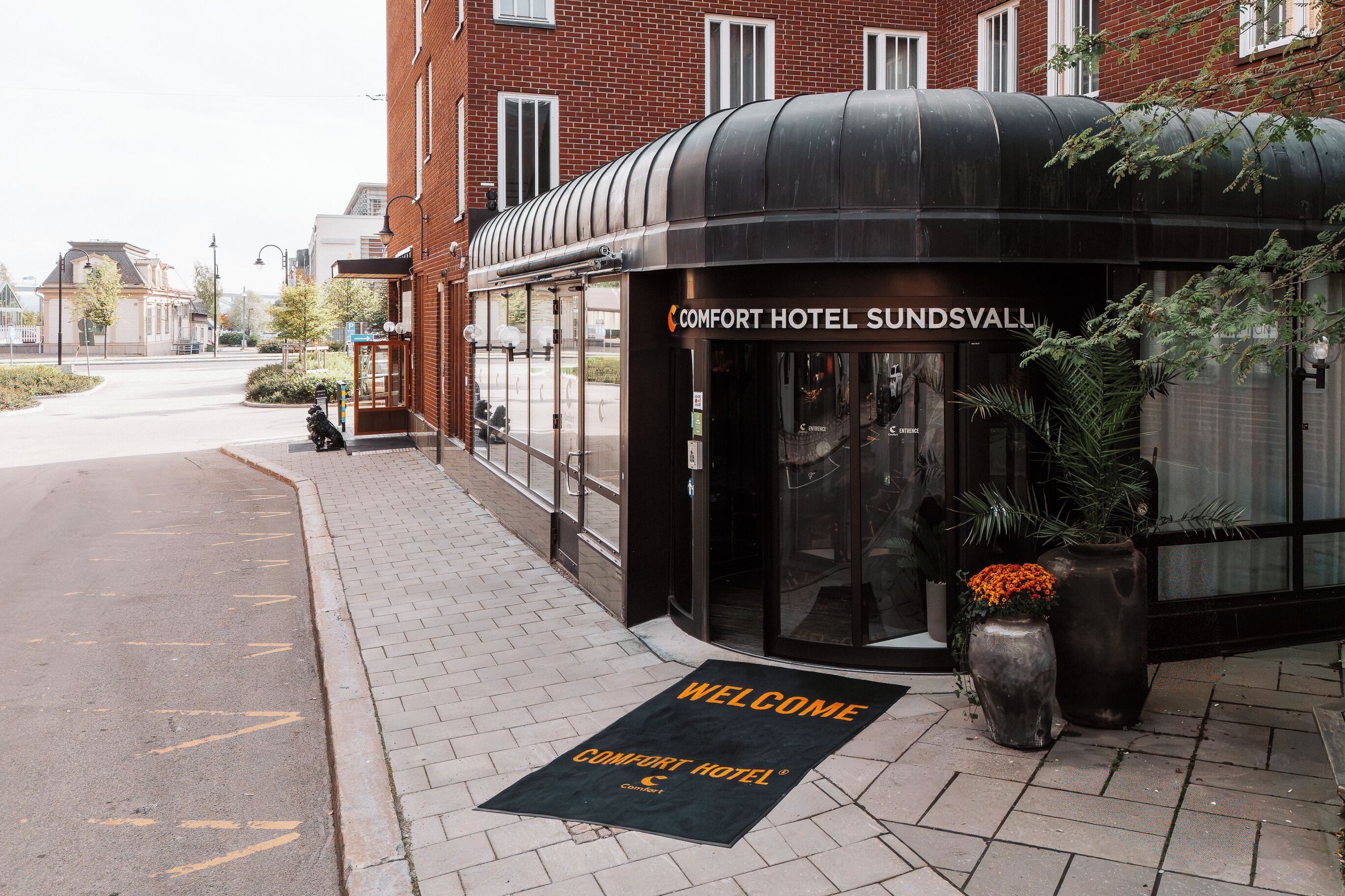 A hotel entrance with a curved black canopy, a welcome mat, and potted plants.