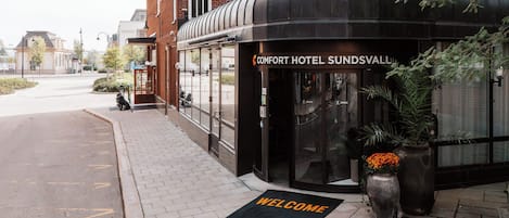 A hotel entrance with a curved black canopy, a welcome mat, and potted plants.