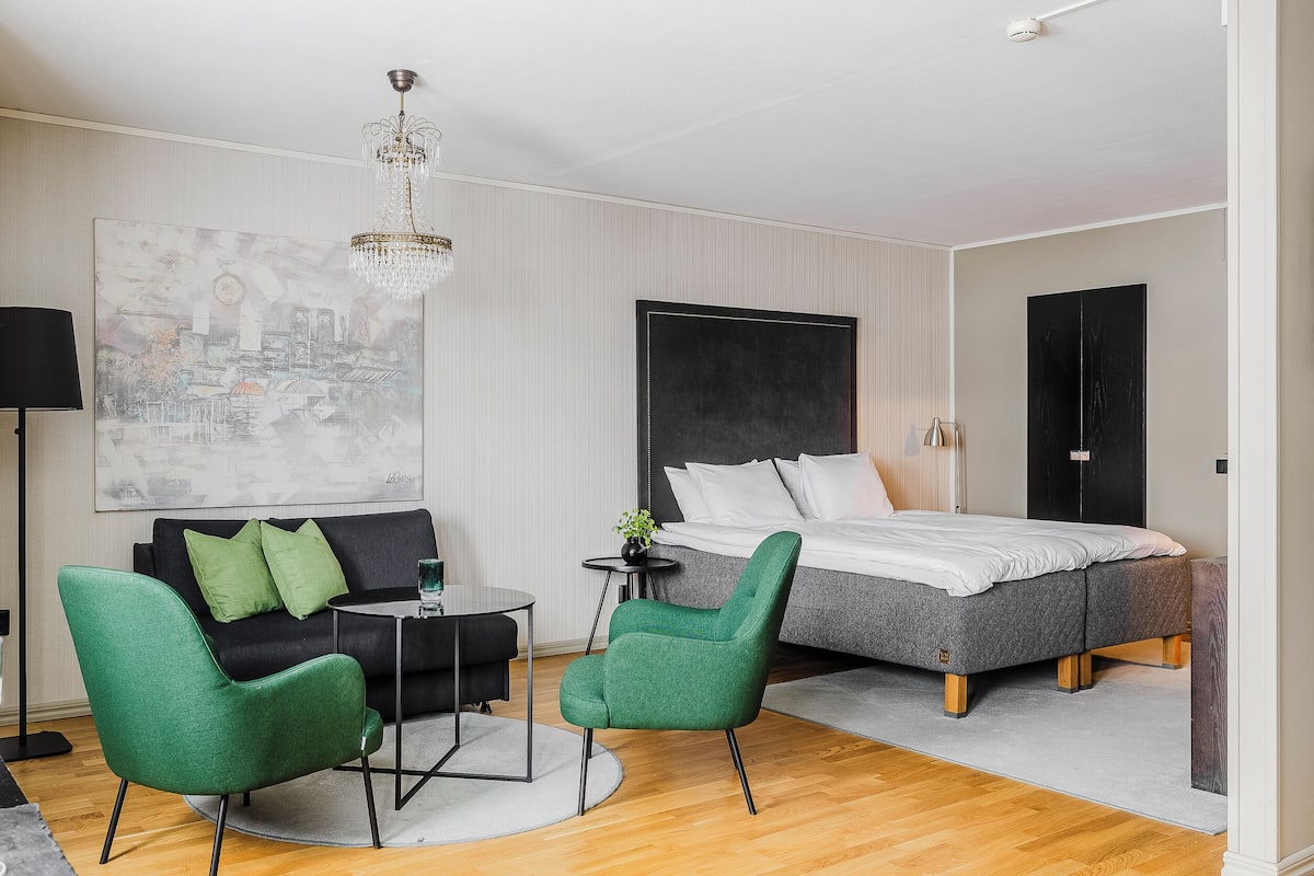 Une chambre d’hôtel moderne avec un lit, un canapé, deux fauteuils verts, une petite table, une lampe sur pied et un grand tableau abstrait accroché au mur.
