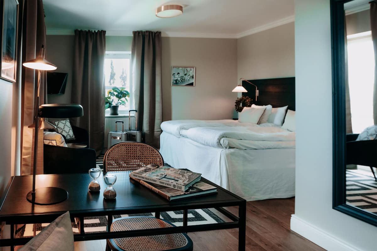 A hotel room with a bed, a desk with a lamp, a chair, a table with books, and a window with curtains.