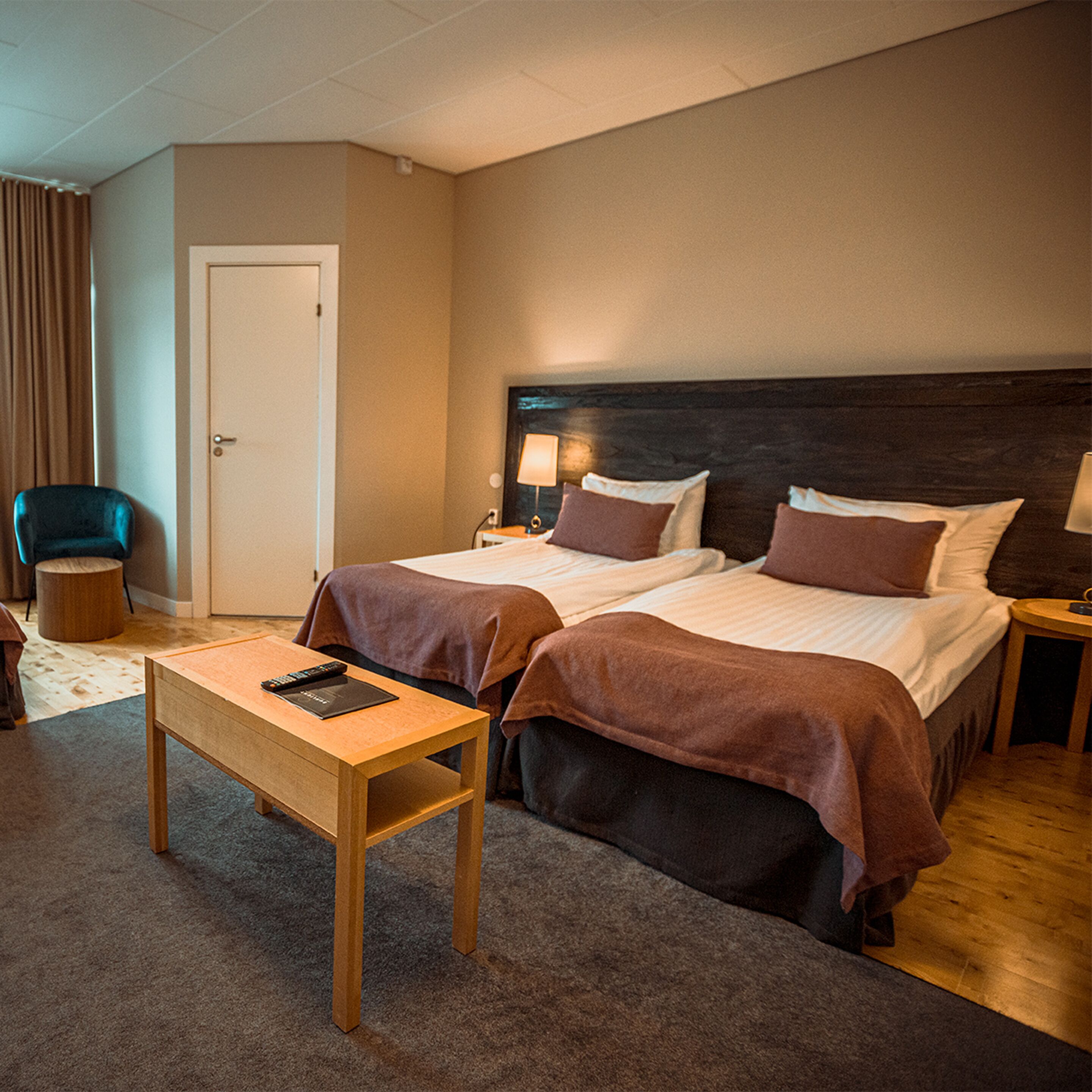 Une chambre d’hôtel avec deux lits, une table en bois, une chaise, une lampe et une porte.