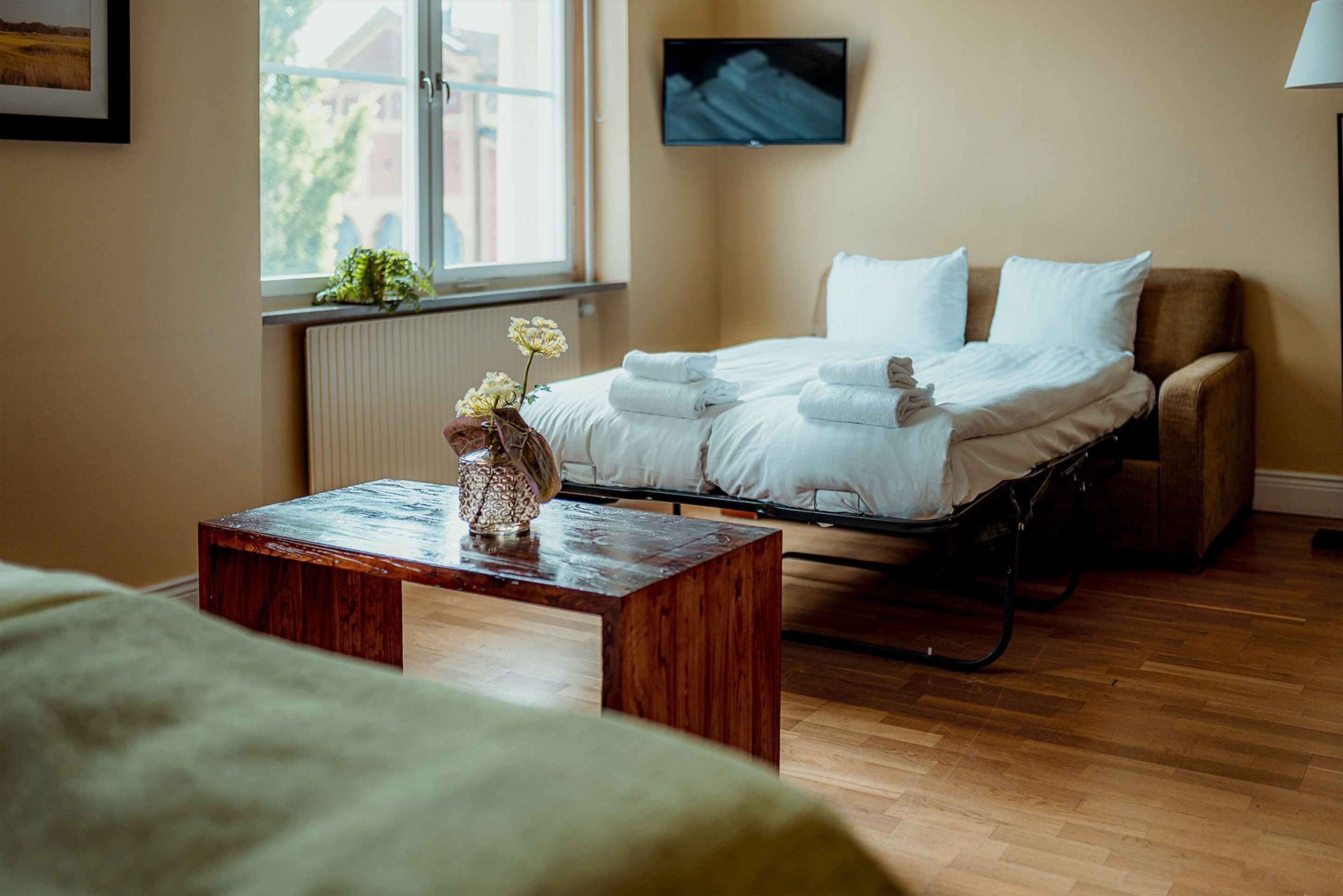 A hotel room with a bed, a wooden table with a vase, a radiator, a window with a view, and a television on the wall.