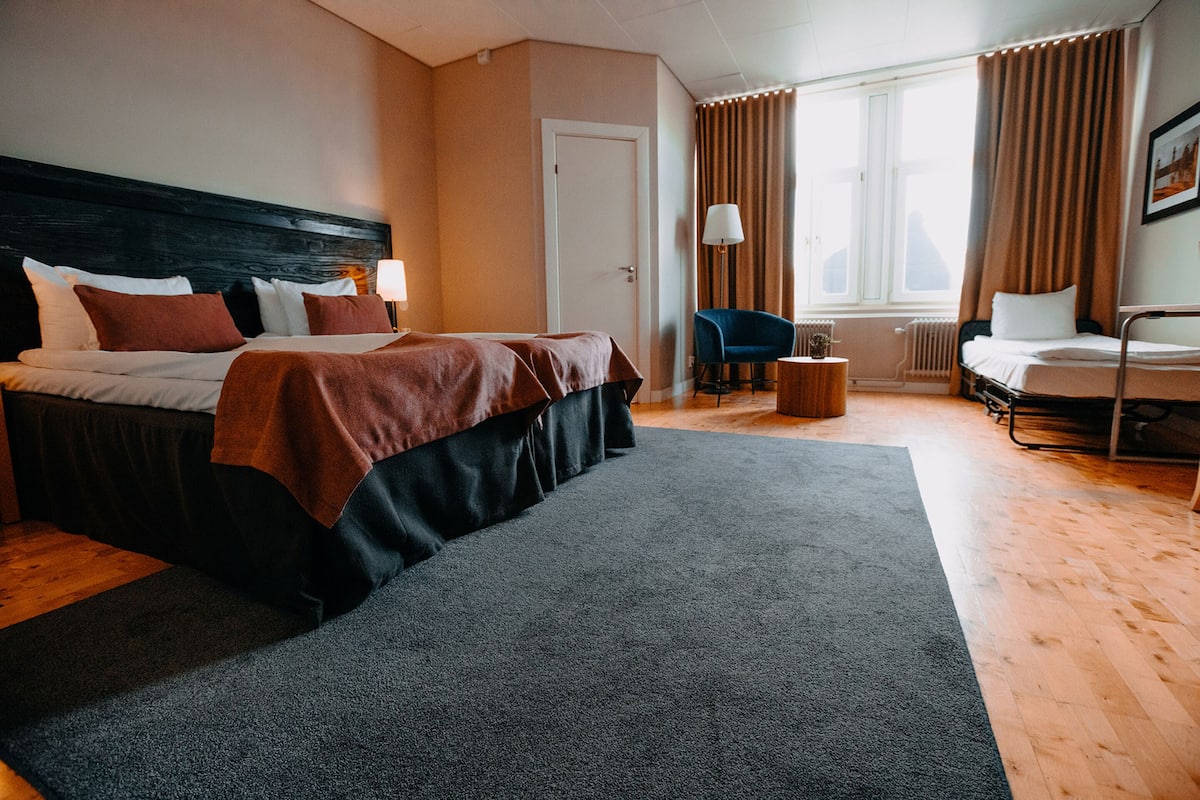 A hotel room with a large bed, a chair, a small table, and a window with curtains.