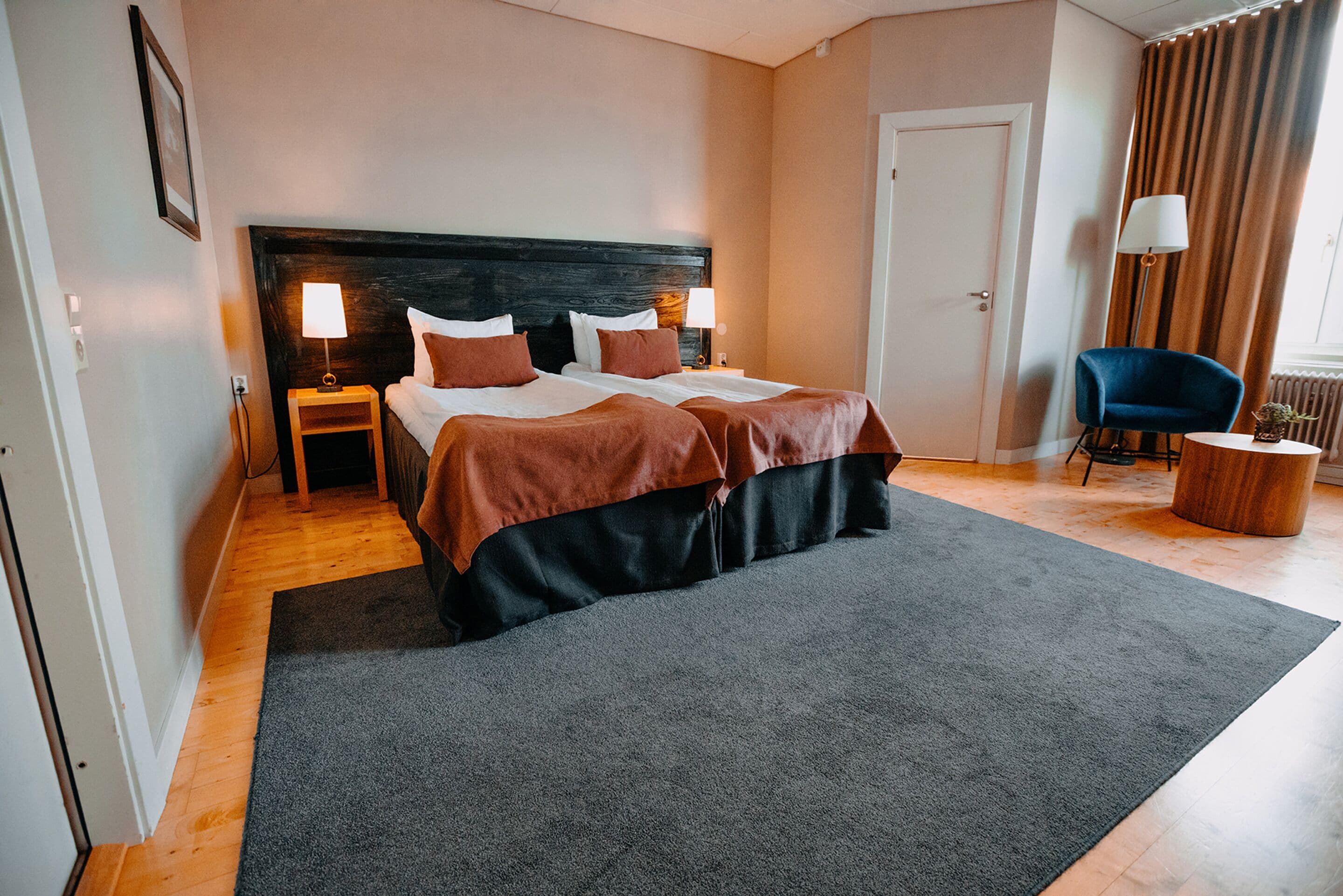 A hotel room with a large bed, two bedside tables with lamps, a blue armchair, and a round wooden side table.
