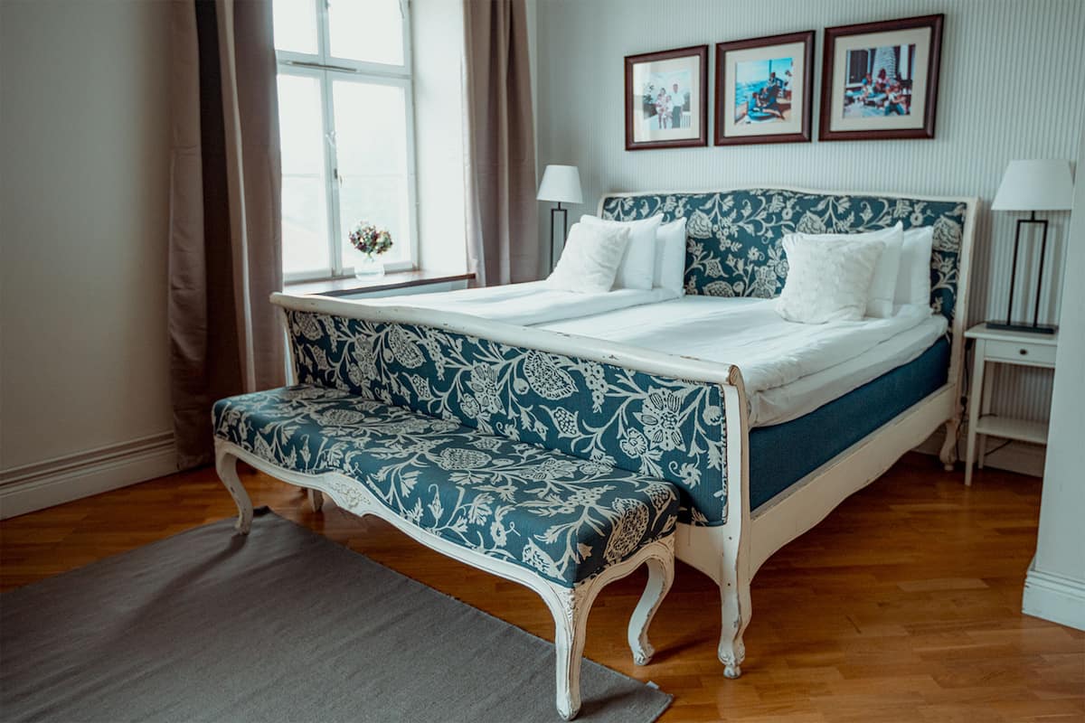 A bedroom with a bed, a bench, a window, and framed pictures on the wall.