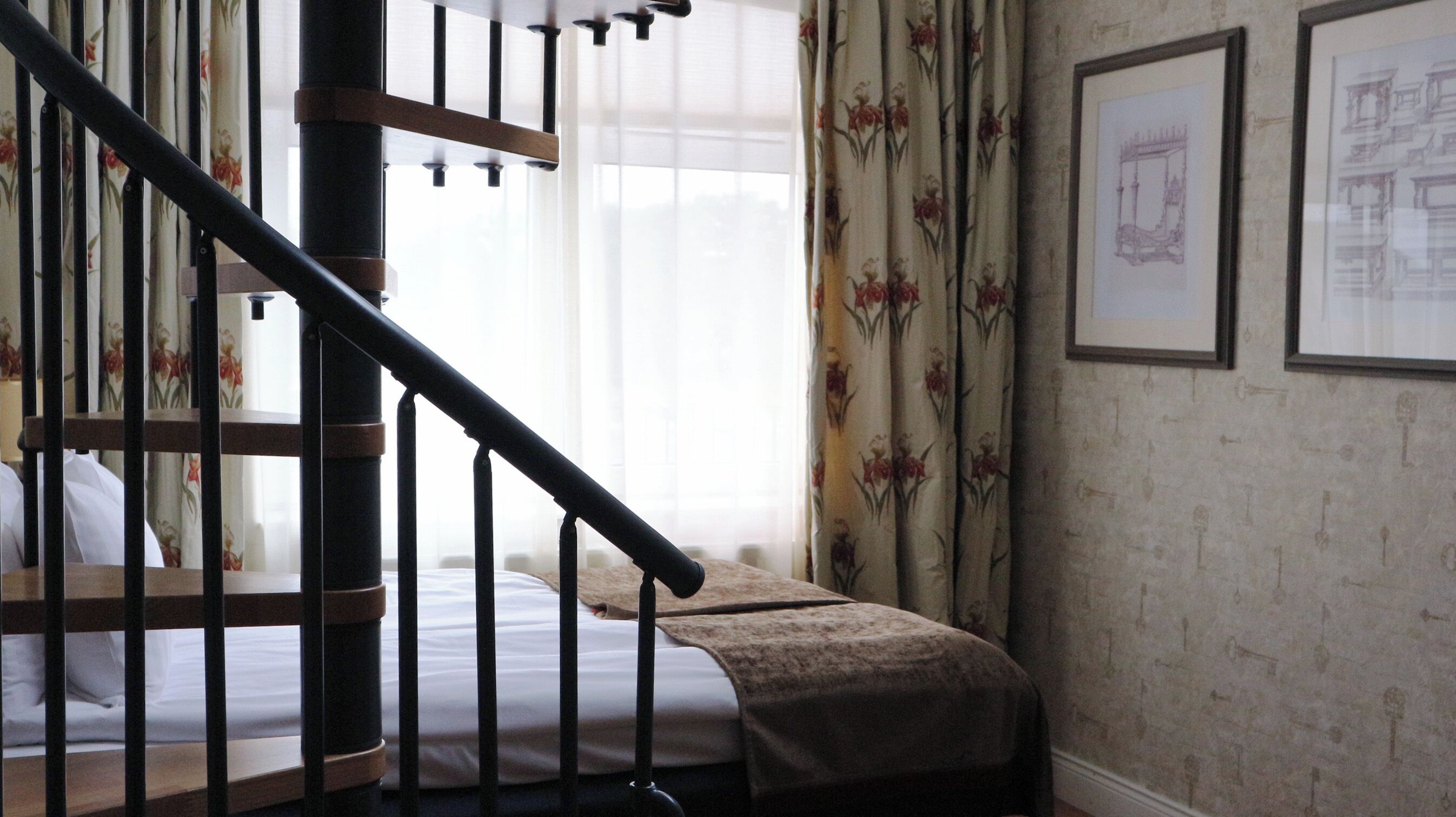 A bedroom with a wooden staircase, a bed with a white mattress, a brown throw blanket, floral curtains, and framed architectural drawings on the wall.