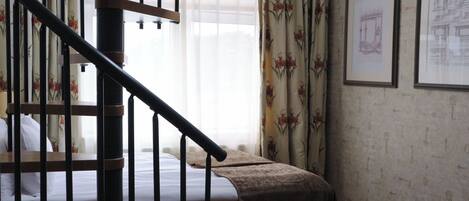 A bedroom with a wooden staircase, a bed with a white mattress, a brown throw blanket, floral curtains, and framed architectural drawings on the wall.