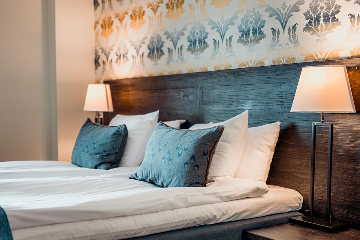 A neatly made bed with a blue headboard, two bedside lamps, and patterned wallpaper.