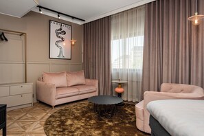 Una habitación de hotel moderna con una decoración en tonos beige y rosa, que incluye un sofá, una mesa redonda, una cama y un amplio ventanal con cortinas.