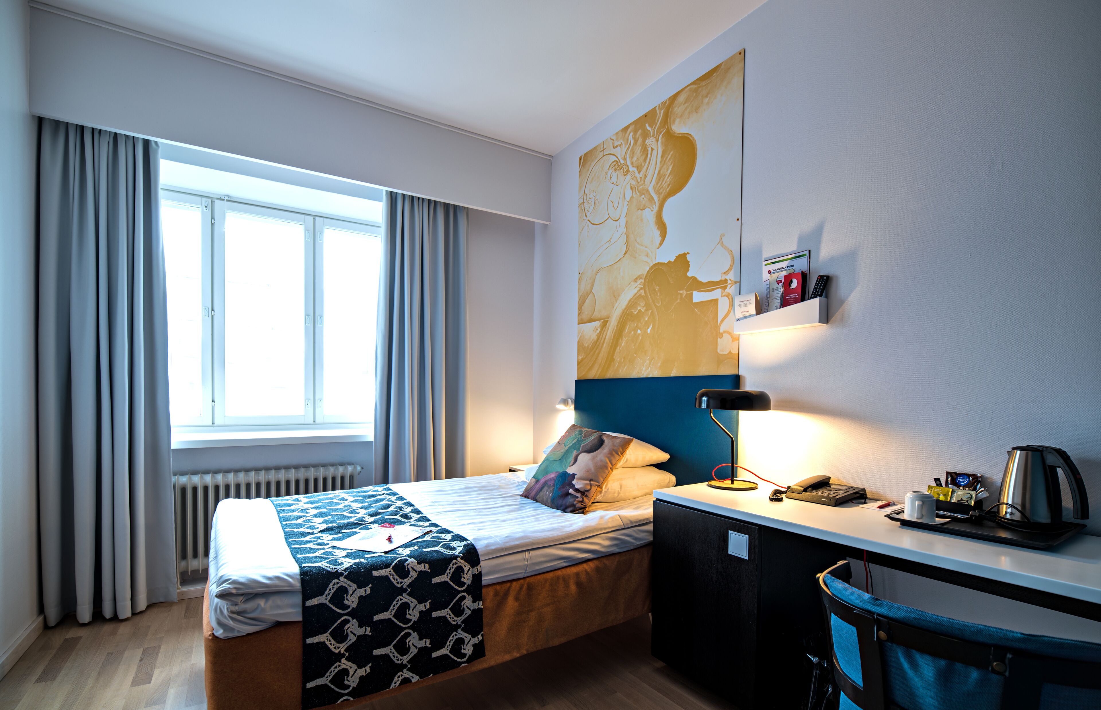 Hotellihuone, jossa on sänky, työpöytä, patteri, ikkuna verhoilla ja suuri abstrakti maalaus seinällä.