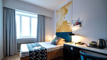 Hotellihuone, jossa on sänky, työpöytä, patteri, ikkuna verhoilla ja suuri abstrakti maalaus seinällä.