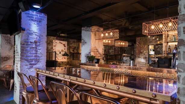Un espace bar doté d’un long comptoir, de chaises en métal et de luminaires suspendus.