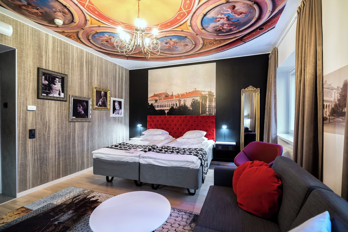 Une chambre d’hôtel moderne dotée d’un grand lit, d’une tête de lit rouge recouverte de tissu, d’un coin salon avec un canapé et des fauteuils, et d’un plafond orné de motifs décoratifs.