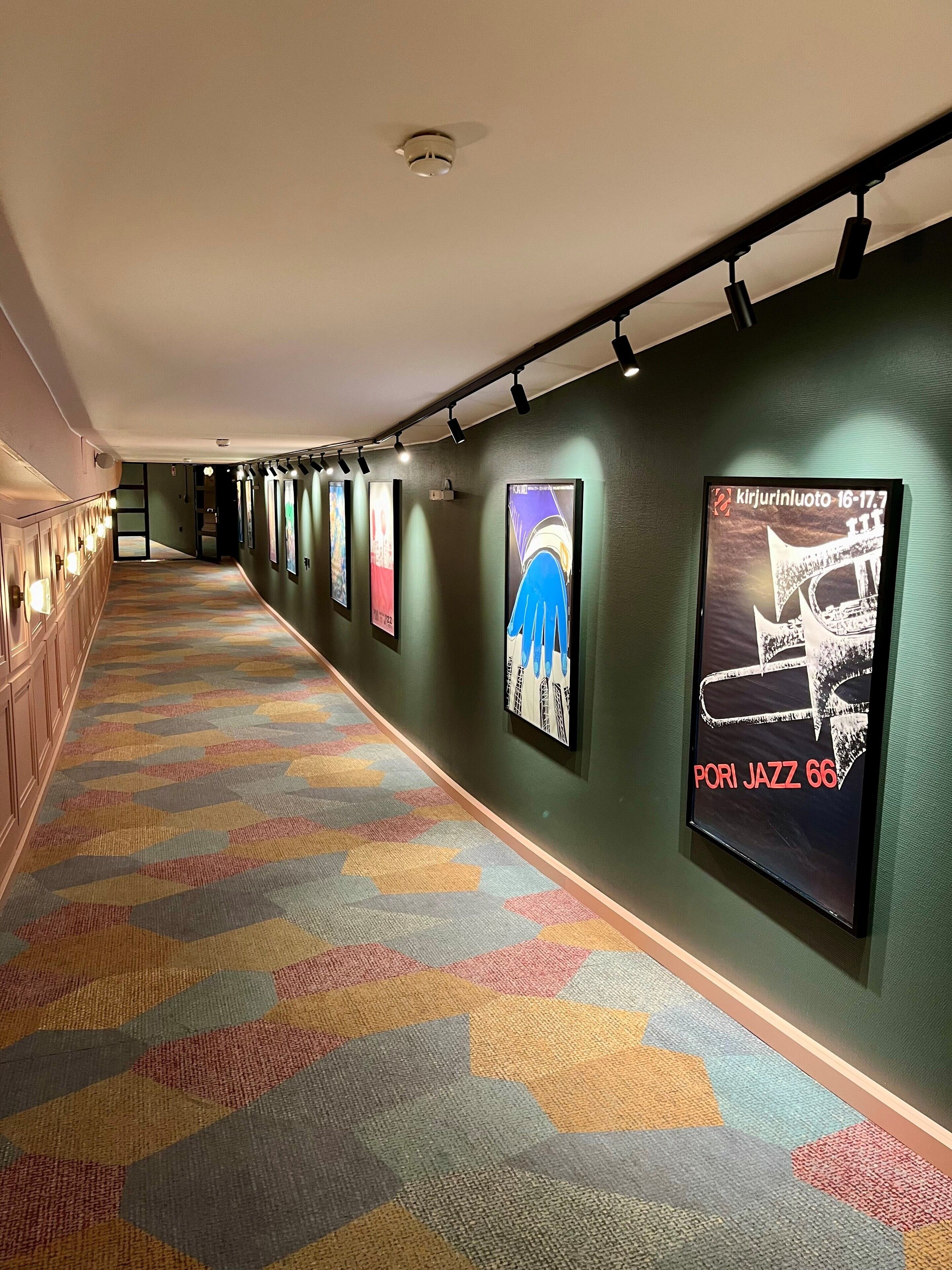 Un pasillo con alfombra estampada, obras de arte colgadas en la pared e iluminación por riel.