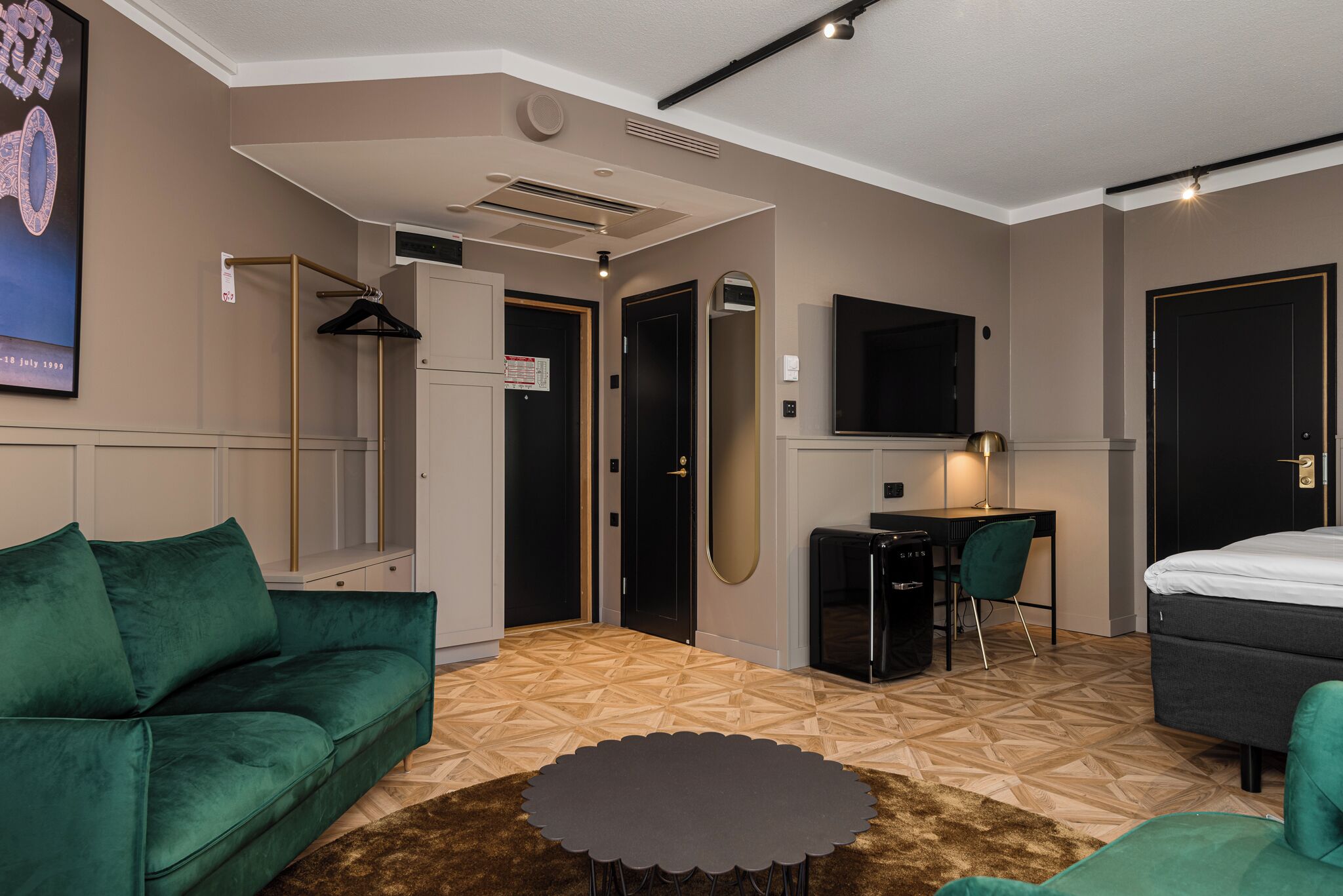A modern hotel room with a green sofa, a small round table, a desk with a chair, a bed, and a TV mounted on the wall.