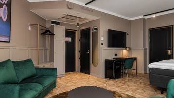 A modern hotel room with a green sofa, a small round table, a desk with a chair, a bed, and a TV mounted on the wall.