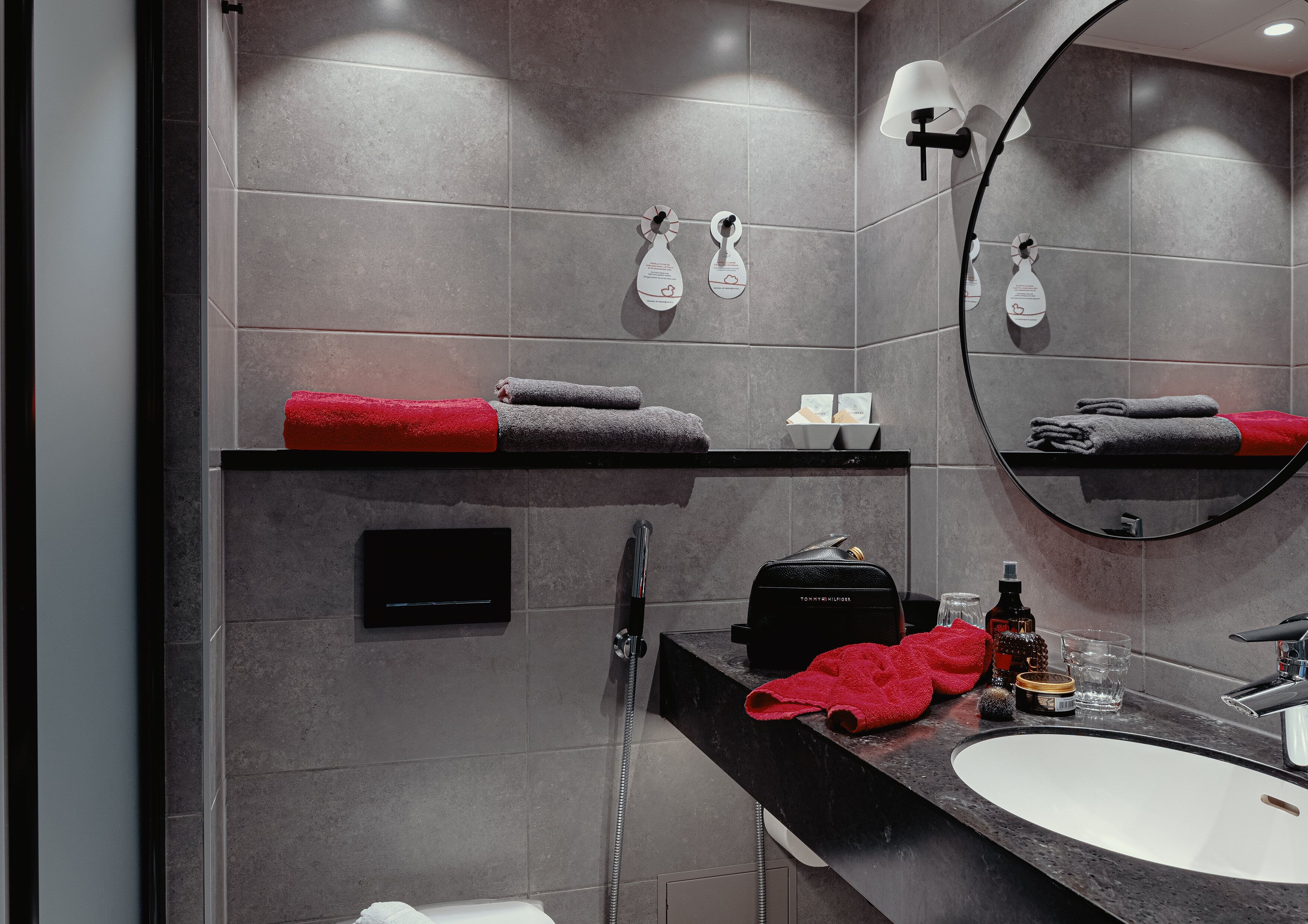 Un baño moderno con baldosas grises, encimera negra, lavamanos, espejo y toallero con toallas rojas.
