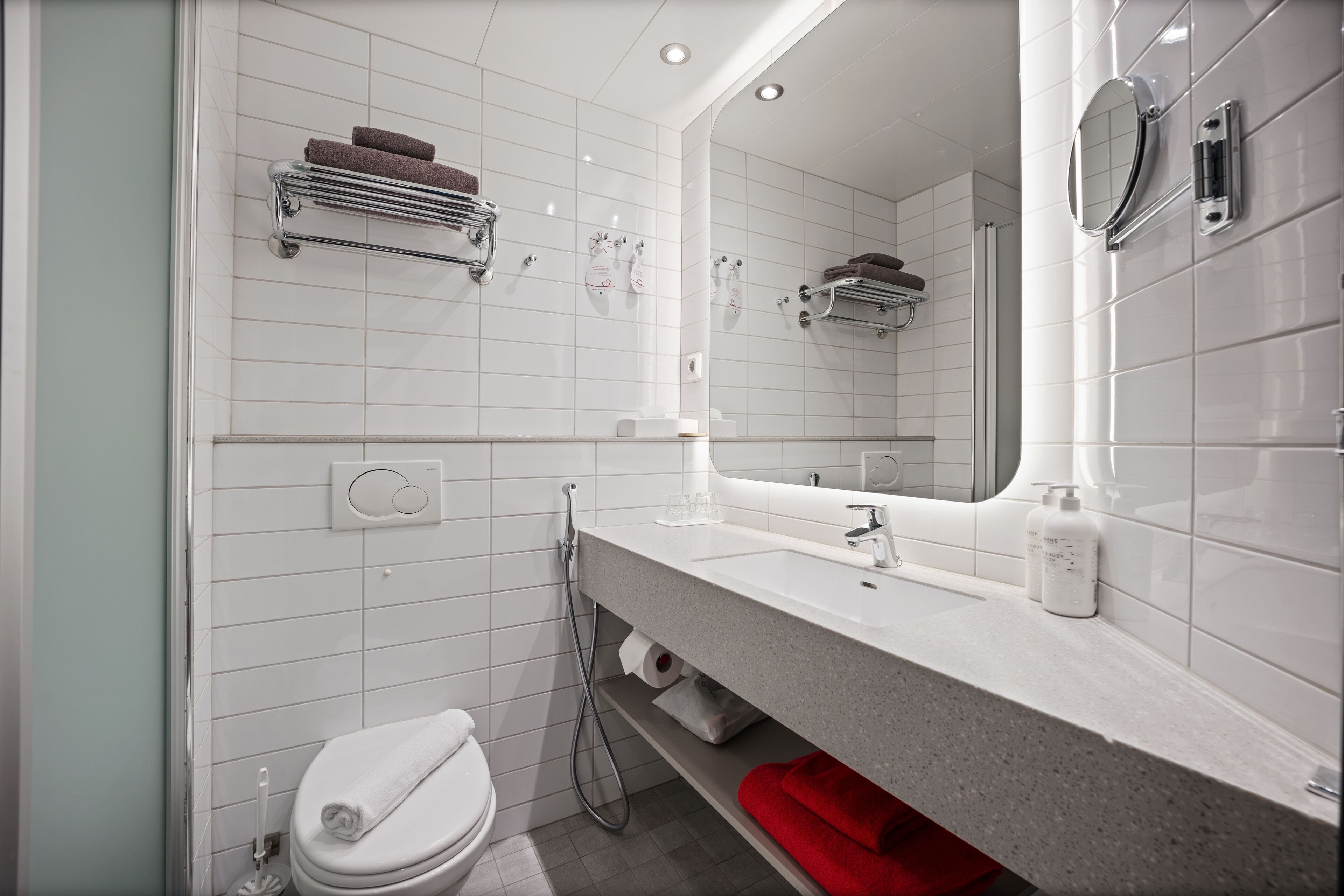 Un baño moderno con pared de azulejos blancos, un espejo grande, una encimera con lavabo y un inodoro.