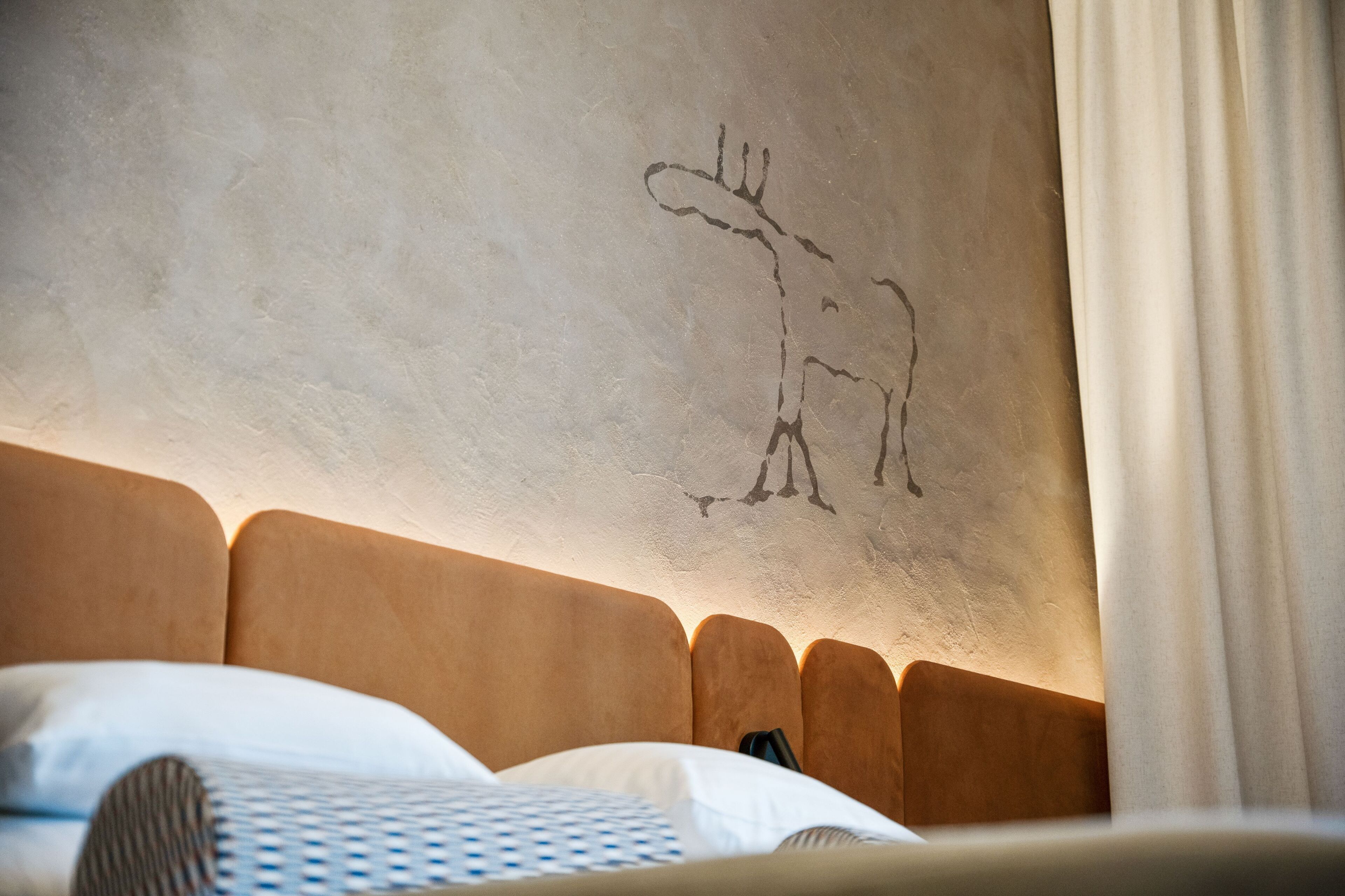 A bed with a headboard, a wall with a simple line drawing, and a curtain.