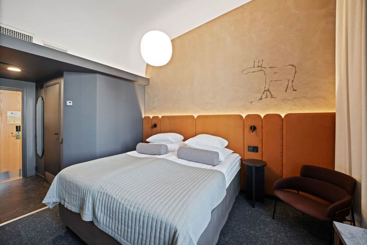 A modern hotel room with a large bed, a small side table, a chair, and a wall with a simple line drawing.