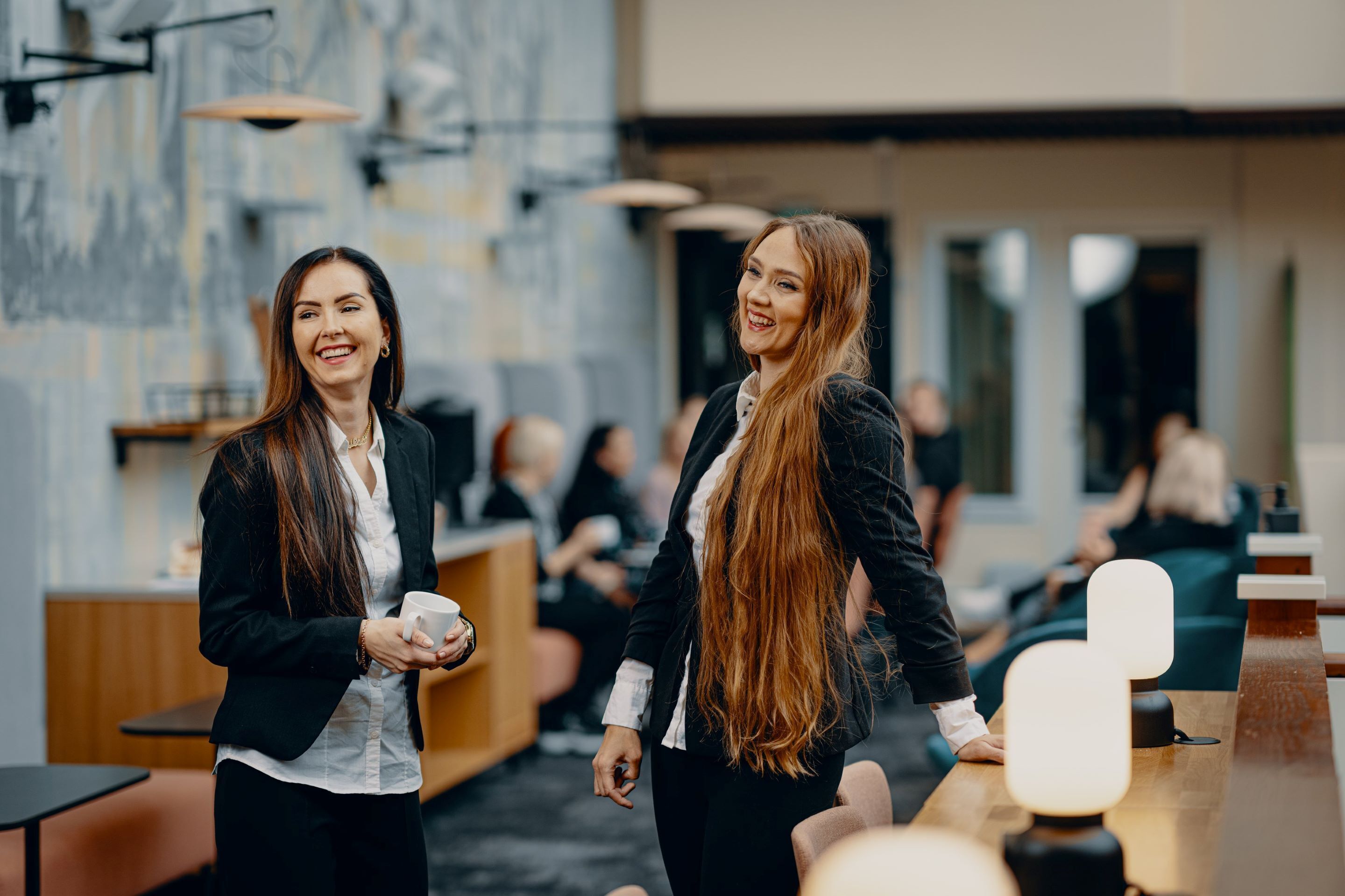 Dos mujeres en un café moderno con mesas y sillas de madera, y una zona de estar informal al fondo.