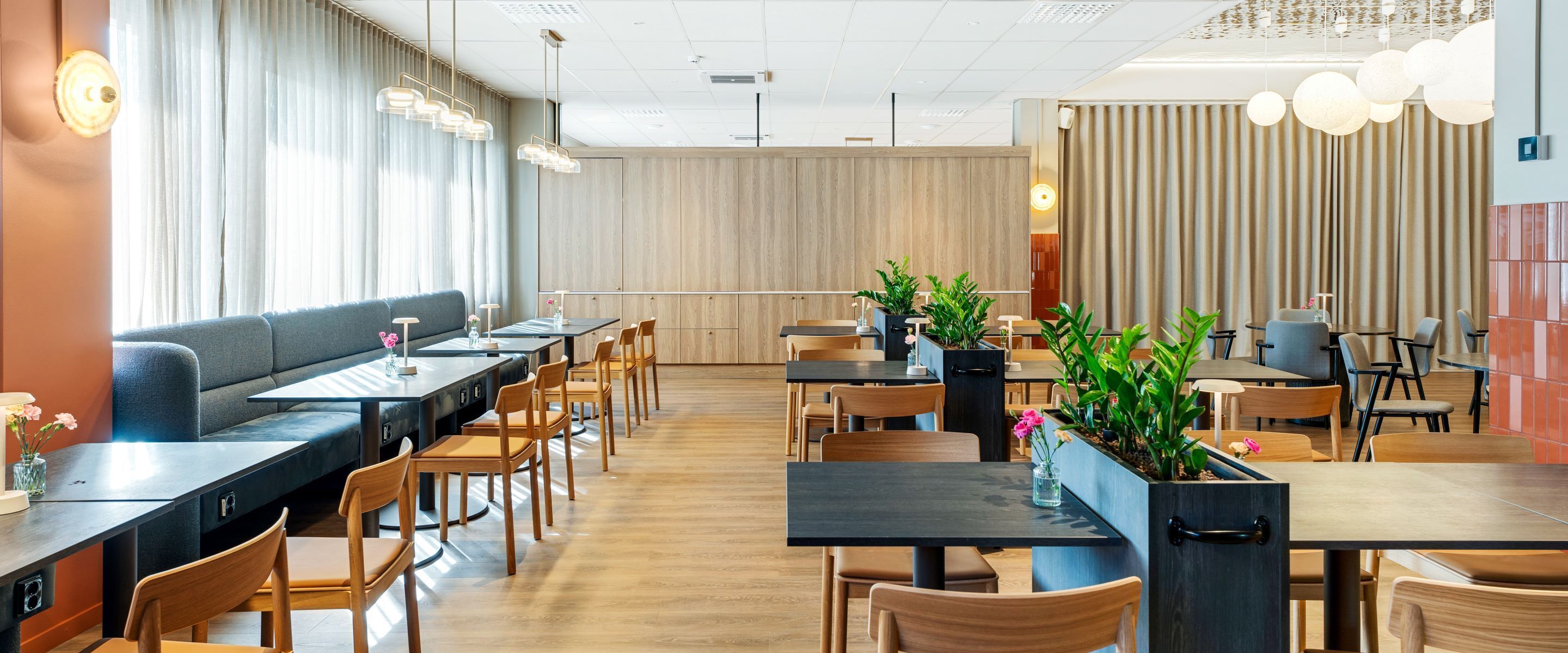 A modern restaurant with wooden flooring, black tables, and wooden chairs. There are potted plants on the tables and a sofa in the background.