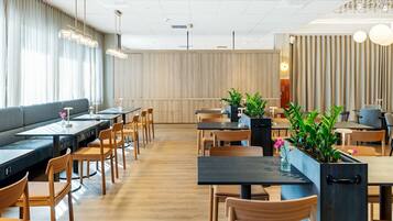 A modern restaurant with wooden flooring, black tables, and wooden chairs. There are potted plants on the tables and a sofa in the background.