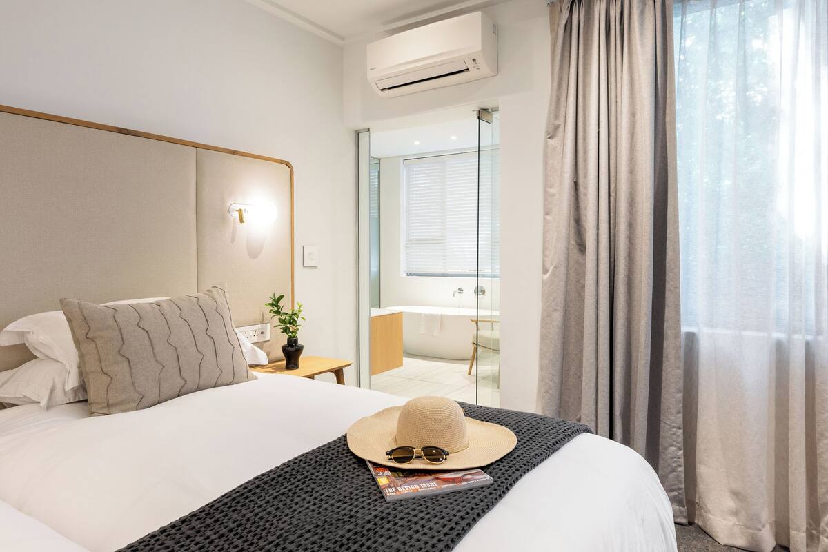 A hotel room with a bed, a nightstand with a lamp, a window with curtains, and an air conditioning unit.