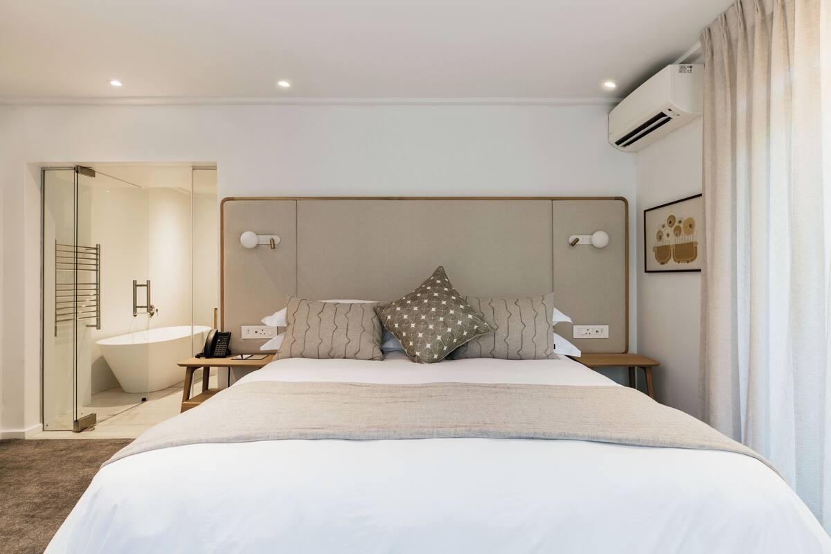 A modern hotel room with a large bed, a bedside table, a nightstand, a built-in bathtub, and a wall-mounted air conditioner.