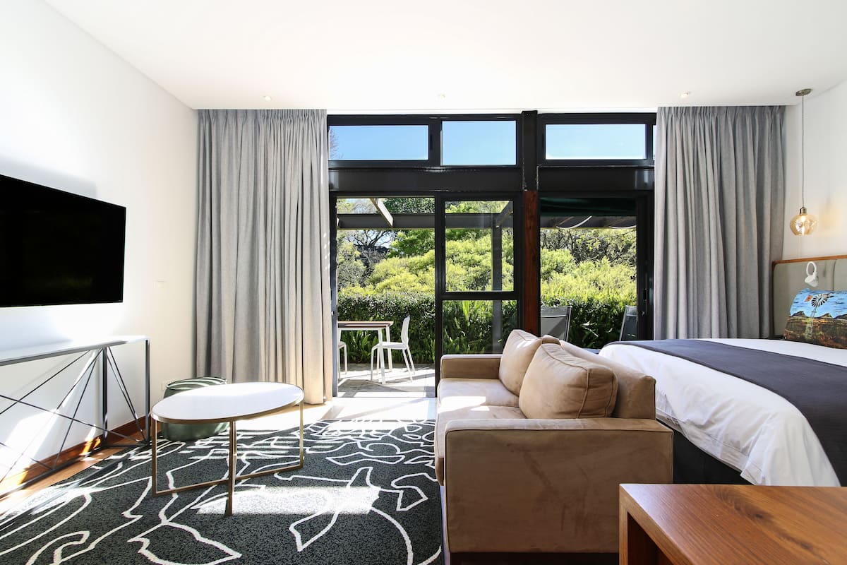 A modern hotel room with a large bed, a sofa, a TV, and a balcony with a view of greenery.